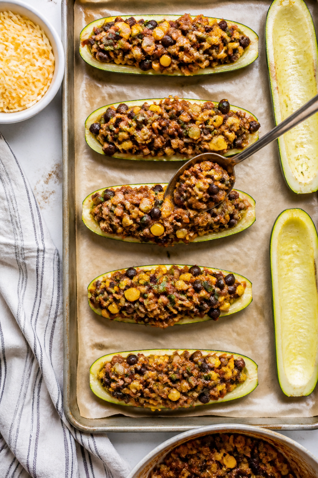 Cooking process: Overhead shot of par-baked zucchini boats being filled on a parchment-lined sheet p
