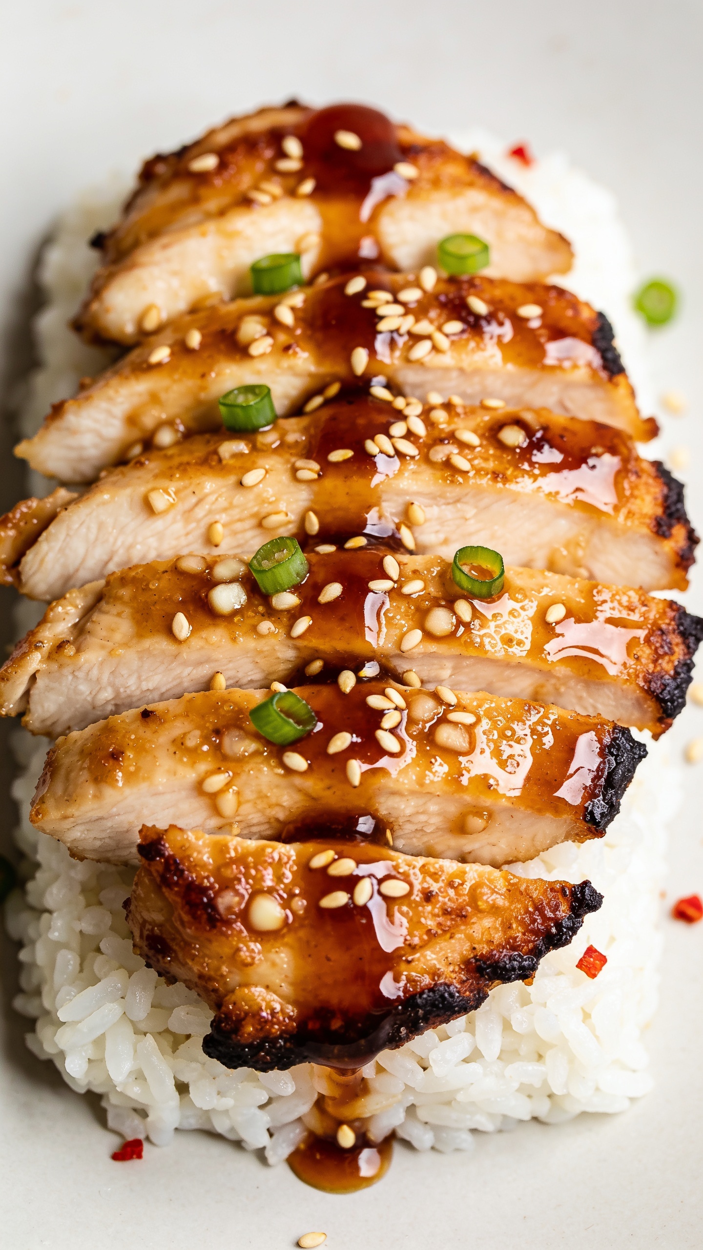 Tight macro angle from slightly overhead of the same honey garlic chicken plated consistently: thick slices fanned over a compact bed of white rice, each piece coated in a thick, reflective honey-garlic-soy glaze with visible minced garlic and micro bubbles of caramelization, edges crisp and dark-golden, a final glossy spoon-drizzle of sauce streaking across the top, toasted sesame seeds and delicate green onion rounds sprinkled evenly, a few flecks of red chili for optional heat; bright natural light, ultra-shallow depth of field to emphasize juicy texture and shine, food fills 90–95% of frame, clean neutral background for premium food-blog styling.