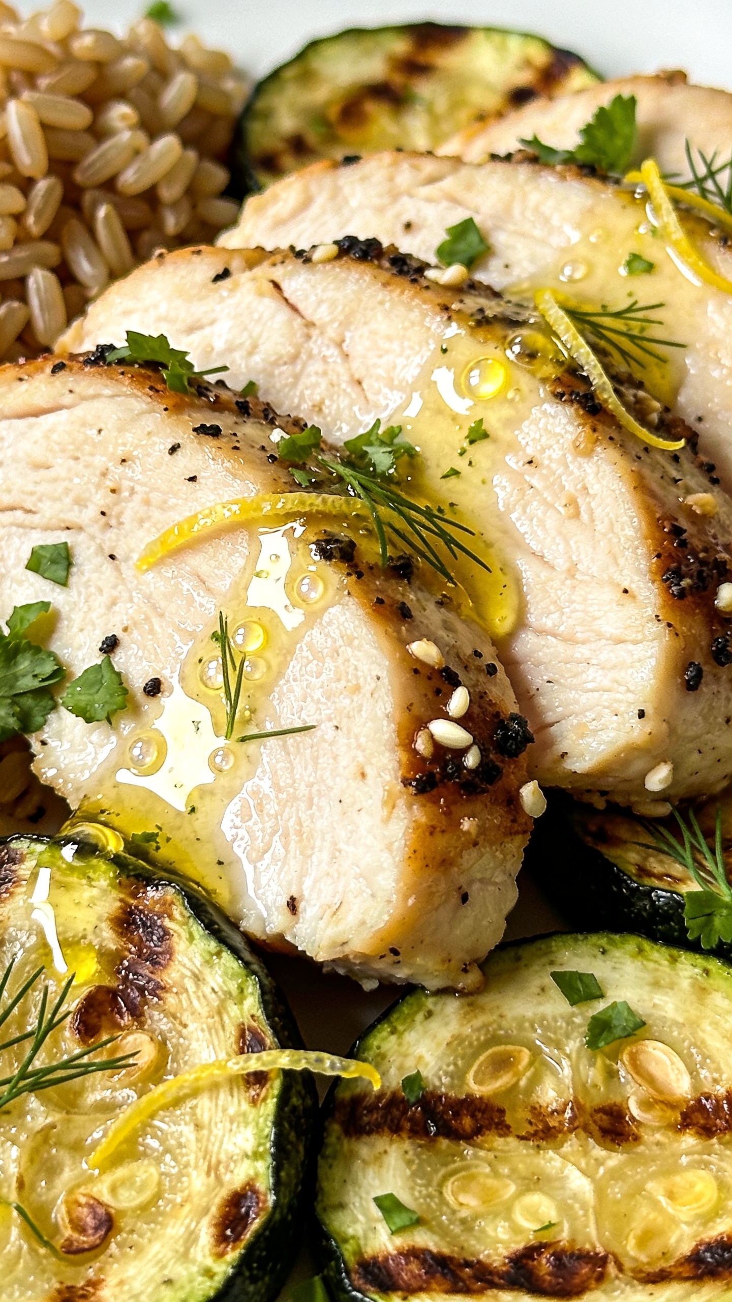 Macro close-up from a low 3/4 angle of the plated meal, emphasizing textures: overlapping slices of juicy seared chicken breast with visible black-pepper and garlic spice crust, nestled against charred zucchini rounds with browned edges and tender centers. A delicate drizzle of lemon-herb olive oil across the top, tiny beads of juice catching the light; fresh chopped parsley and dill sprinkled, fine lemon zest strands glowing. A compact portion of brown rice peeks in the back corner, grains distinct. Bright natural side light, creamy bokeh, food filling 90–95% of frame, saturated colors, crisp highlights, cohesive with the other images.