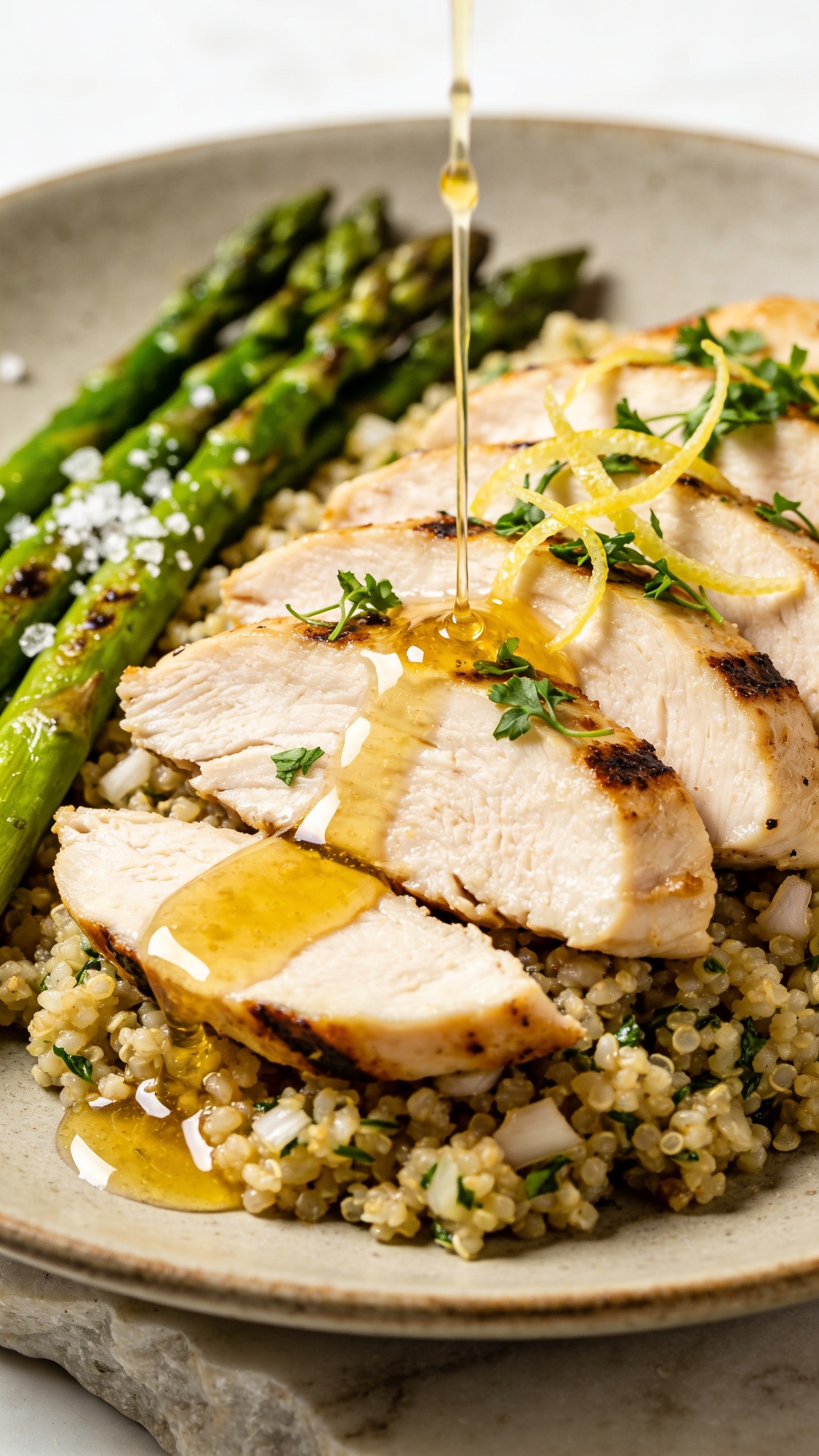 Extreme close-up from a lower angle focusing on texture: sliced chicken breast fanned over garlic-herb quinoa with seared edges and a lacquered lemon-Dijon-honey glaze pooling lightly; a thin stream of extra glaze just finished drizzling, forming glossy droplets; asparagus spears aligned alongside, vibrant green with light blistering and coarse sea salt crystals; micro parsley sprinkled across chicken and quinoa, lemon zest strands catching the light; bright natural lighting, shallow depth of field isolating the front slice of chicken; shot on a neutral ceramic plate over pale stone, food occupying 90â95% of the frame, premium editorial finish, no text.