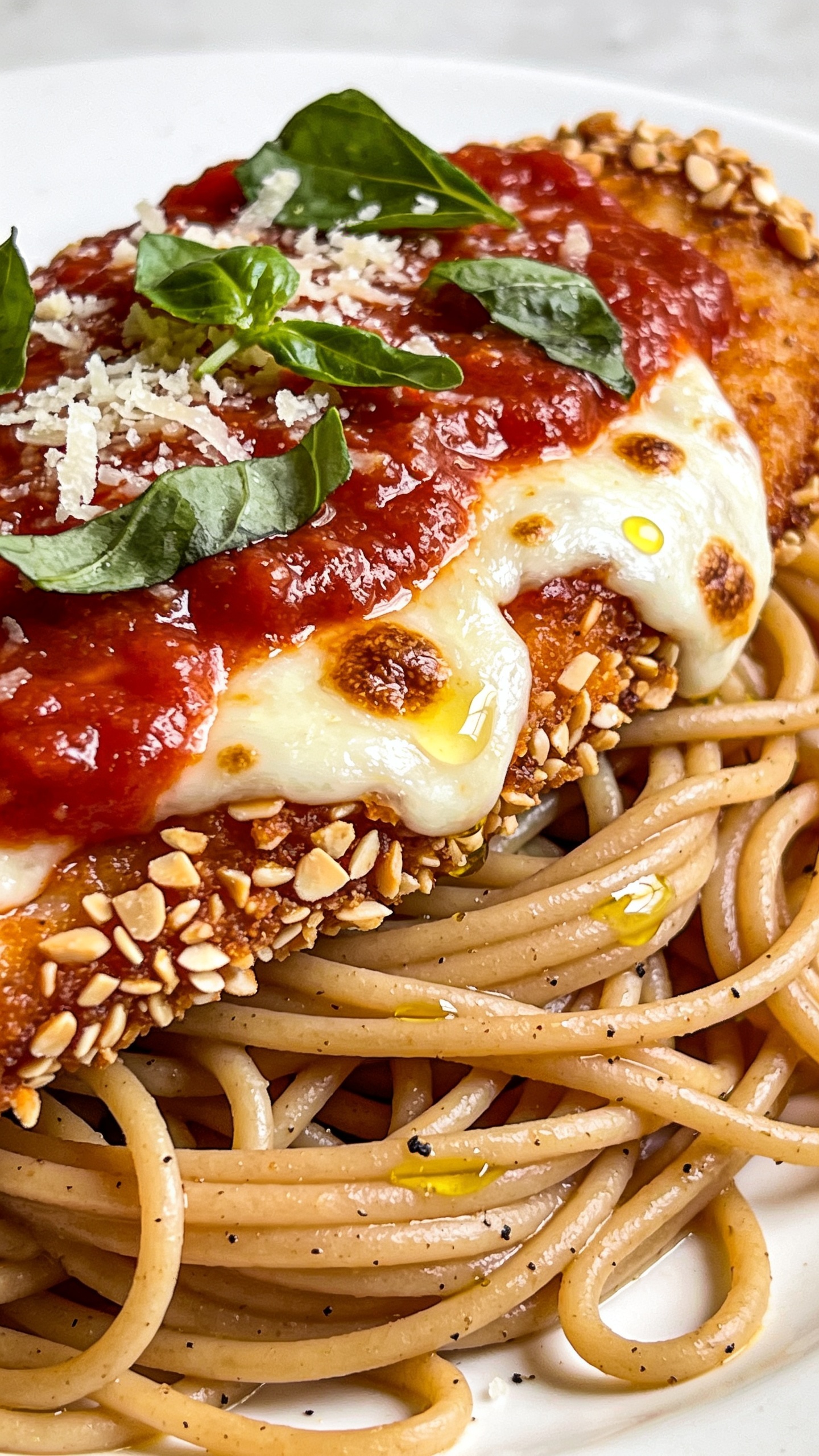 Extreme close-up angled from the side to showcase textures: the same finished Chicken Parmesan resting on a light swirl of whole-wheat spaghetti—jagged, crunchy almond-crumb crust along the cutlet’s edge; marinara pooled and glistening; light mozzarella melted with browned spots and tiny oil beads; freshly grated Parmesan scattered with torn basil leaves on top. A faint sheen of olive oil on pasta strands, coarse black pepper visible. Bright natural backlighting, shallow depth of field, tight crop with food occupying 90–95% of the frame, rich colors and crisp details.