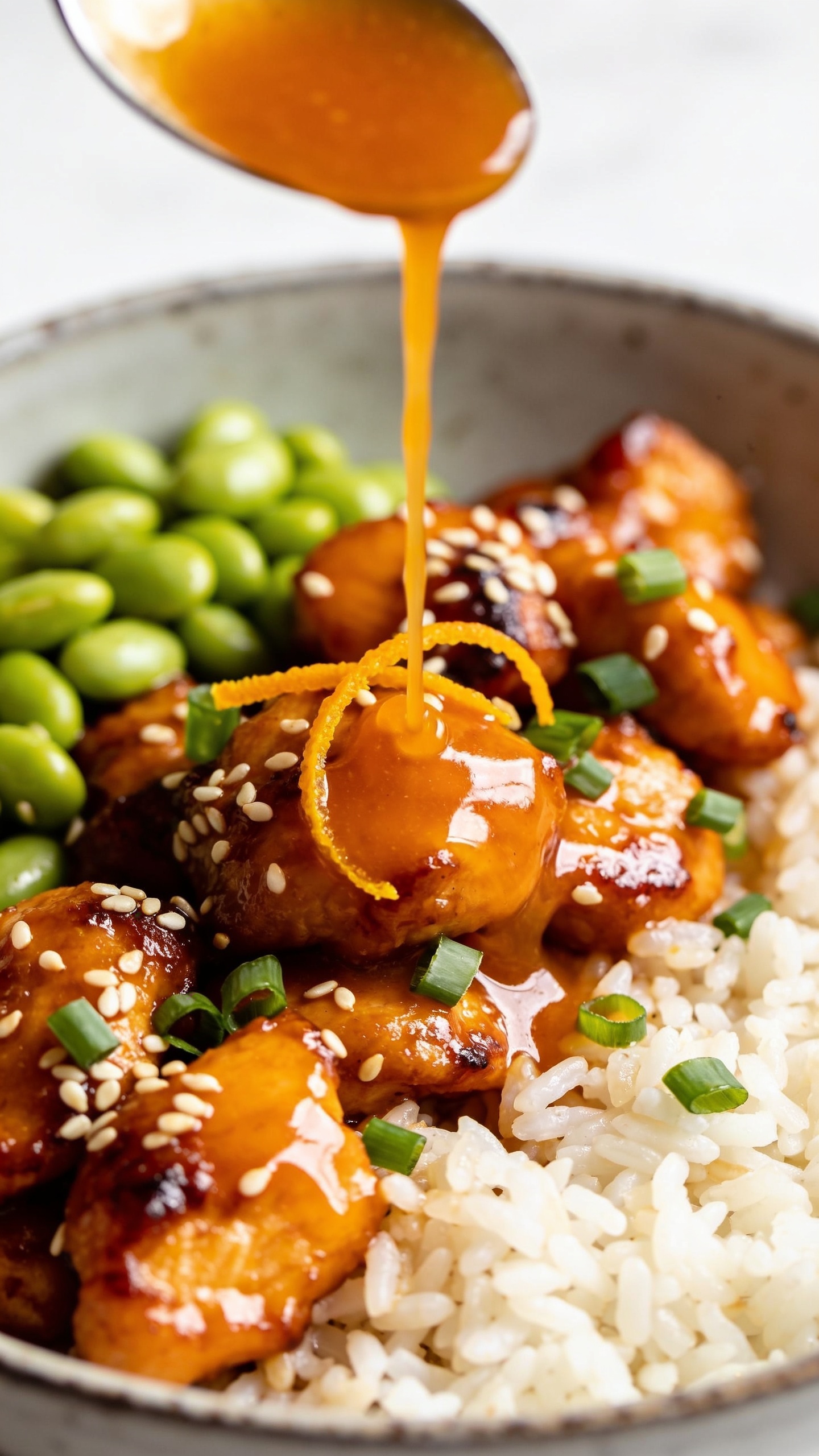 Tight overhead-angled close-up focusing on texture: a spoonful of extra orange glaze being slowly drizzled over the same orange chicken and jasmine rice bowl; the sauce forms glossy highlights across the caramelized edges of the chicken, sesame seeds and scallion greens catching the light; edamame pods clustered on one side, a small arc of fresh orange zest scattered over the chicken; shallow depth of field blurs the far edge of the bowl, bright natural light, minimal props, 90–95% frame filled with richly colored, irresistible food.