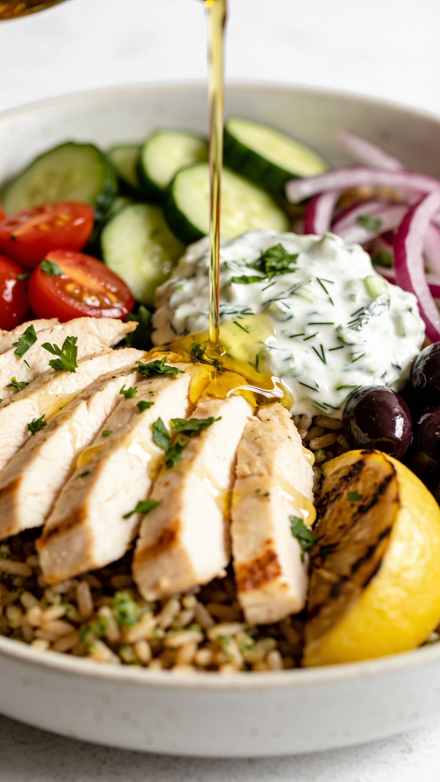 3. Tight macro angle from the opposite side of the same finished bowl: chicken slices fanned across lemon-herb brown rice, tzatziki dolloped with visible grated cucumber and dill, a slow drizzle of golden olive oil captured mid-pour over the chicken and veggies, creating glossy highlights; vibrant cucumbers, cherry tomatoes, red onion, Kalamata olives, and a charred lemon wedge tucked in; finishing sprinkle of chopped fresh parsley; bright natural lighting, shallow depth of field, 90–95% food in frame, same plating, textures, and color palette as images 1–