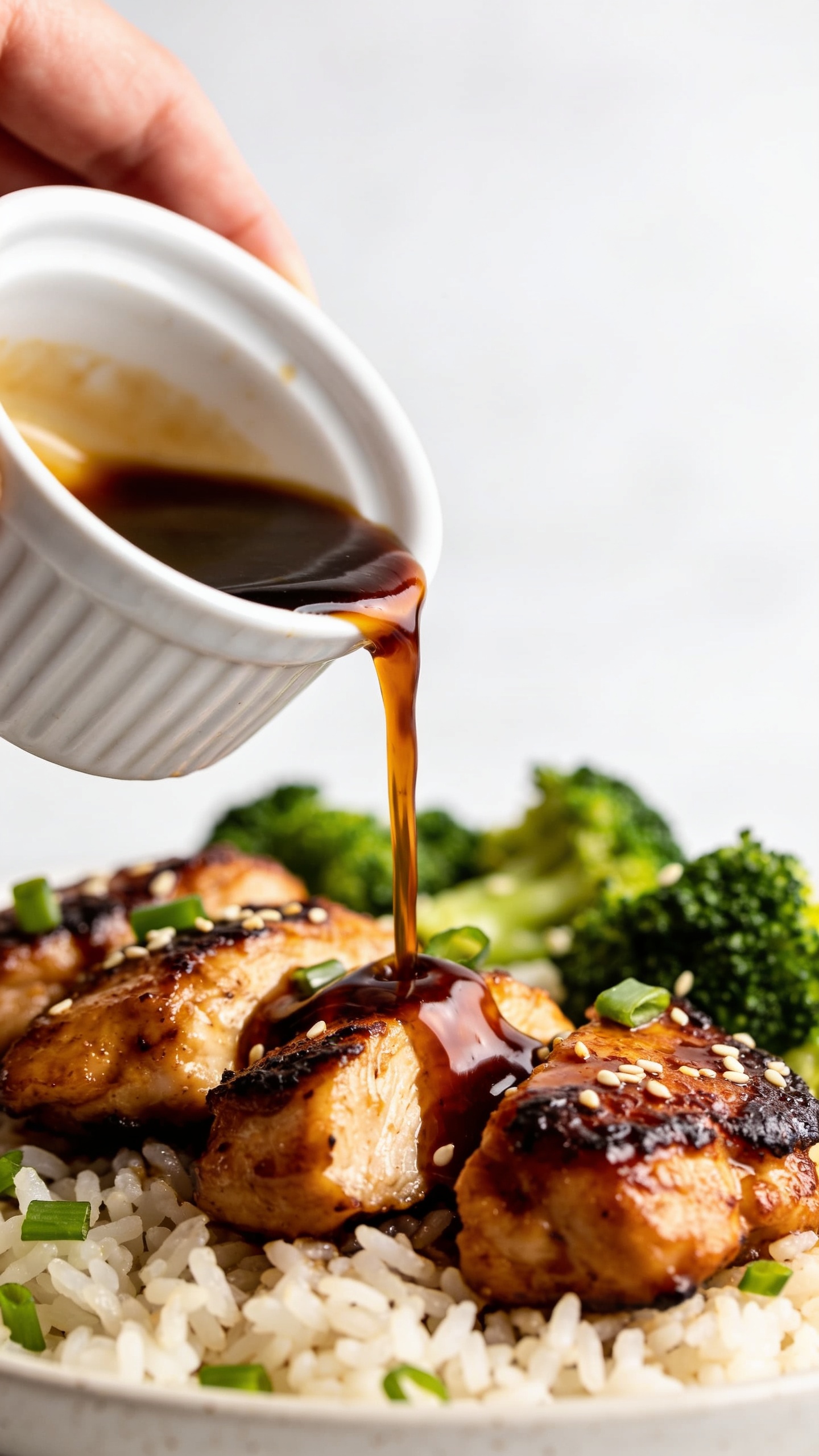 Ultra-close detail shot from a low 3/4 angle of the same finished recipe: bourbon chicken pieces nestled over jasmine rice with broccoli, a slow ribbon of extra bourbon glaze being poured from a small spouted ramekin onto the chicken, freezing mid-pour as it pools and cascades between pieces. Surface highlights emphasize sticky thickness; crispy caramelized edges and juicy interiors visible. Fresh green onions and sesame seeds scattered naturally. Bright natural lighting, shallow depth of field, food dominating 90–95% of the frame, cohesive with the first images, clean neutral backdrop.