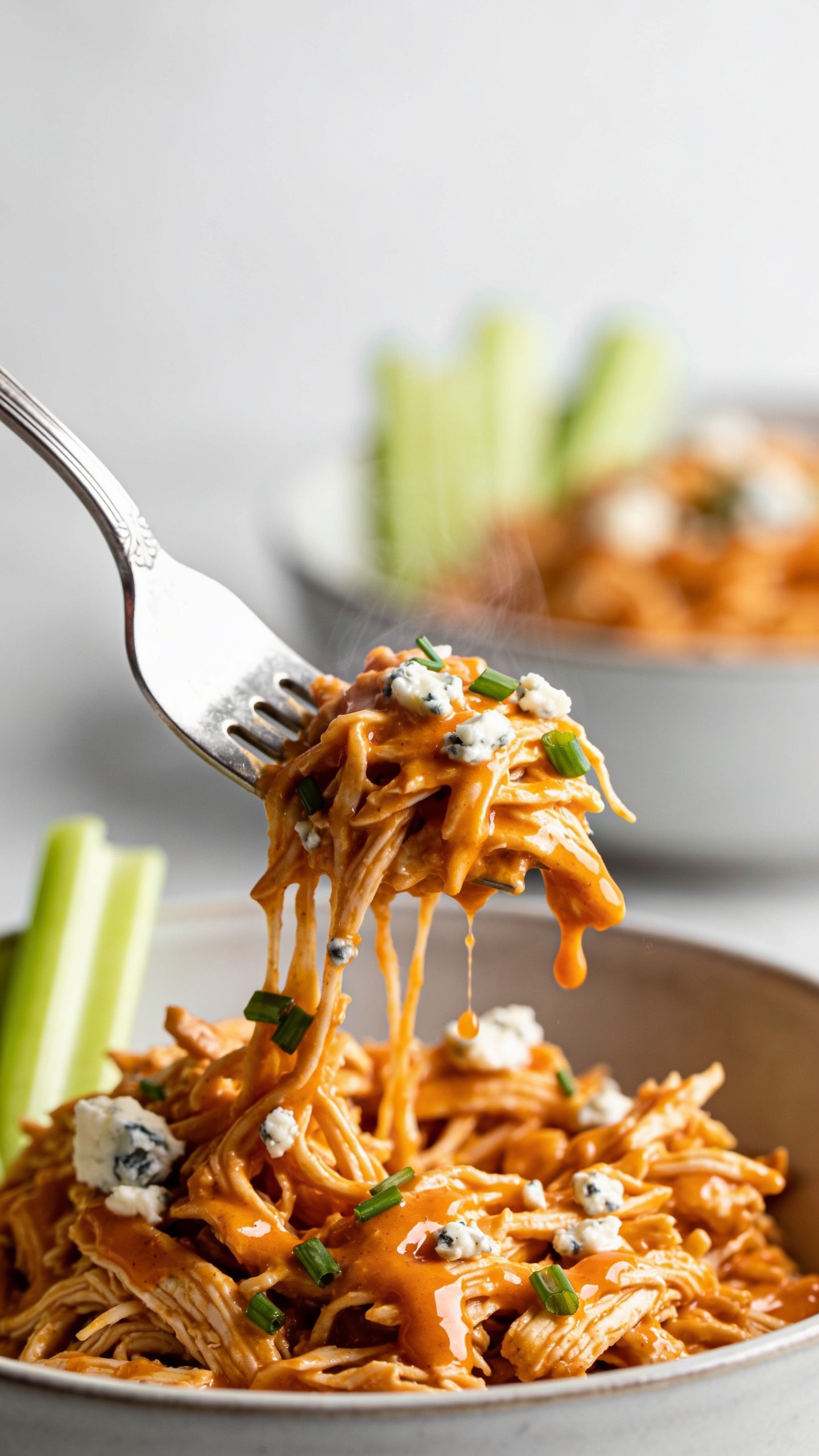 Dynamic action close-up capturing the perfect bite: a silver fork lifting a small, juicy mound of the same shredded buffalo chicken directly above the bowl, strings of saucy chicken pulling from the pile, droplets of glossy orange buffalo sauce clinging and about to drip; faint steam rising; tiny crumbles of blue cheese speckling the bite, with a few chopped chives stuck to the sauced strands. Background softly blurred but clearly the same bowl of buffalo chicken with celery accents. Bright natural light, shallow depth of field, 90–95% food in frame, no hands, only the fork visible, movement frozen one second before the bite.