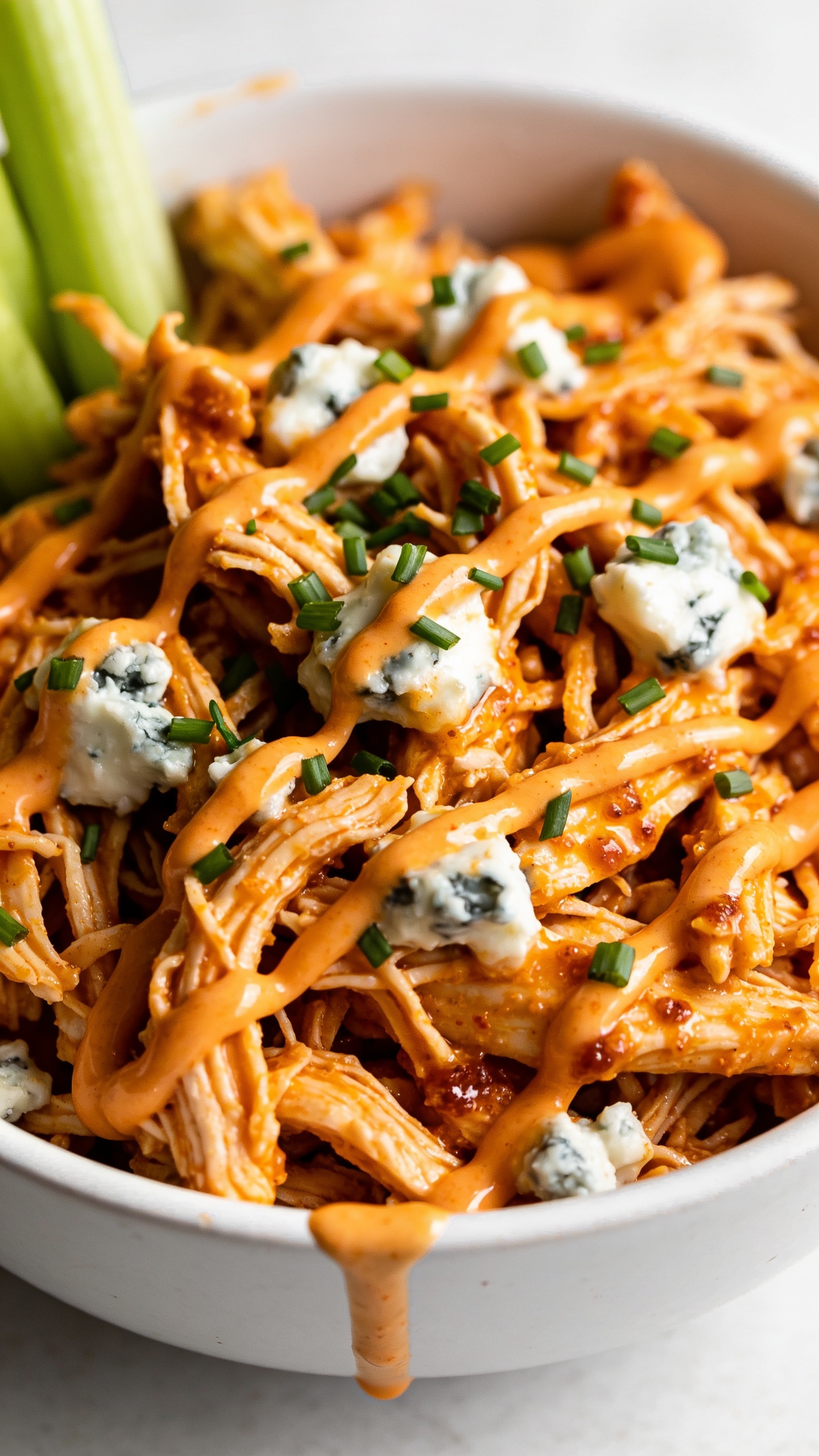 Tight macro overhead-angled close-up of the same finished buffalo chicken in the same white bowl: succulent shreds interwoven and lacquered in vibrant orange sauce, micro-beads of fat glistening, edges slightly crisped from finishing under the broiler, scattered blue cheese crumbles melting into warm pockets, fine shower of freshly chopped chives across the top; a light extra drizzle of buffalo sauce zigzagging over the surface with a small pool at the rim, celery sticks peeking in at the margin. Bright natural light, shallow depth of field, 90–95% of frame filled with the chicken, rich textures and vivid color, clean neutral background.