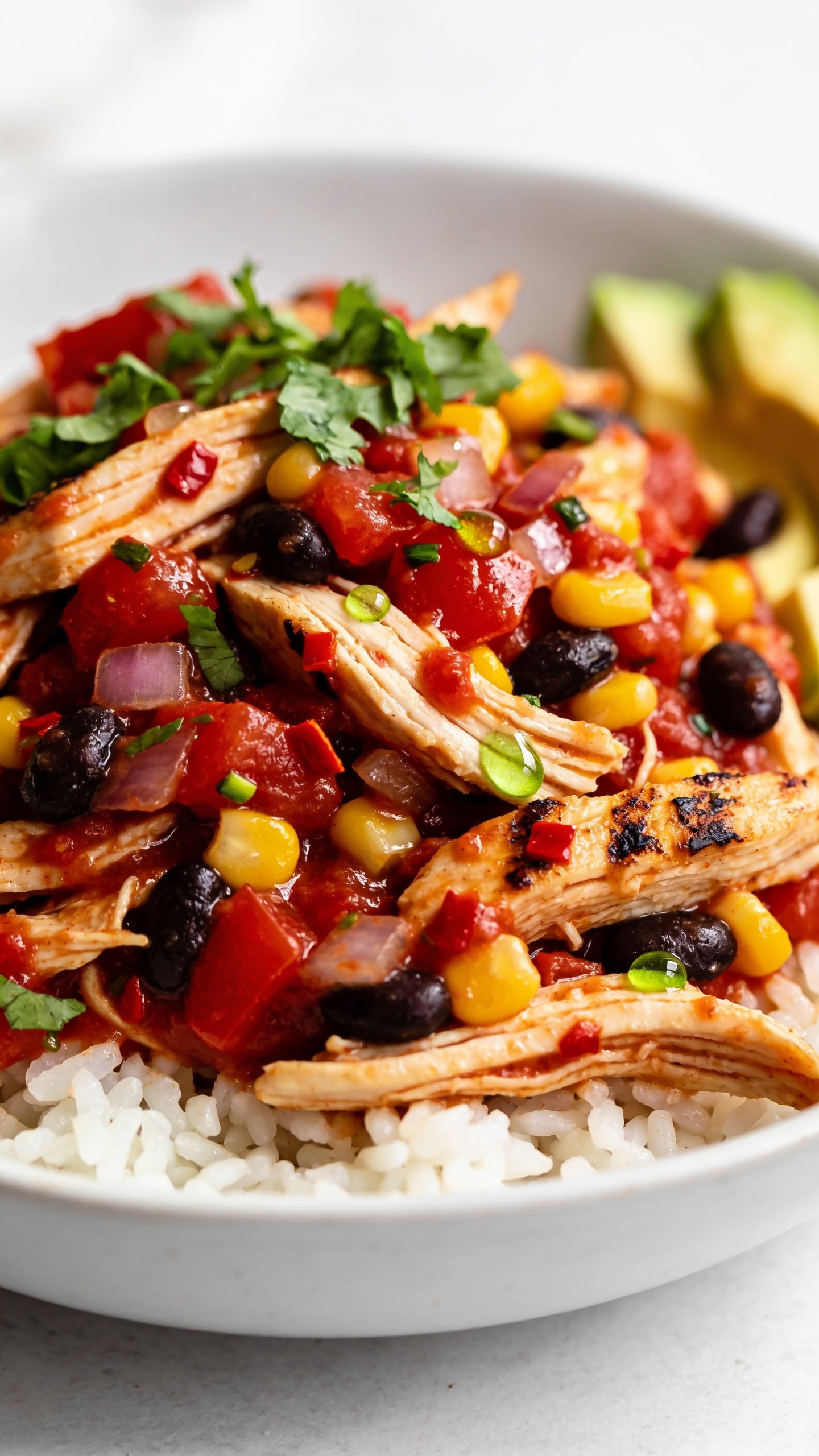 Extreme close-up hero shot of High Protein Crockpot Salsa Chicken: juicy shredded boneless skinless chicken breast tossed in chunky red salsa with visible tomato, onion, and chili flecks, mixed with black beans and sweet corn kernels for added protein and color. The chicken is glossy and moist, lightly charred edges from broiler finish, garnished with fresh chopped cilantro and a squeeze of lime that leaves tiny droplets glistening on the surface. Served in a shallow wide white bowl over a small bed of fluffy white rice, with a few avocado slices tucked to the side but mostly obscured by the saucy chicken. Bright natural side lighting, shallow depth of field, vibrant reds, yellows, and greens, premium food-blog styling, 90–95% of frame filled with food, no text, no hands.