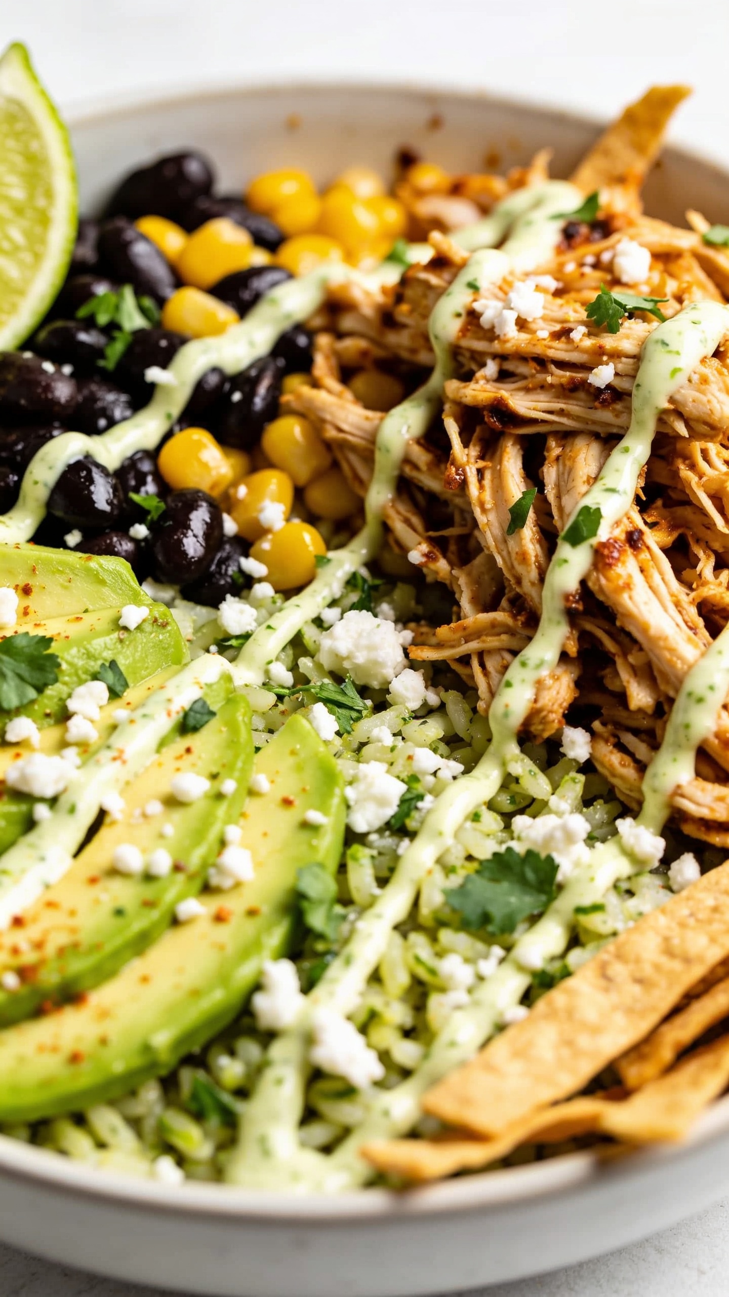 Extreme close-up side angle of the same finished burrito bowl emphasizing juicy textures and glossy sauces. Focus on the shredded chicken piled against cilantro-lime rice, with shiny black beans and corn nestled in; chipotle-lime crema drizzled in a fine zigzag, catching light; crumbled cotija and fresh cilantro scattered; avocado slices fanned and lightly speckled with chili-lime seasoning; lime wedge edge visible. Crisp tortilla strips add texture near the rim. Bright natural lighting, shallow depth of field isolating the chicken and crema, saturated but true-to-life colors, 90–95% frame filled with food, matching the hero’s styling and ingredients.