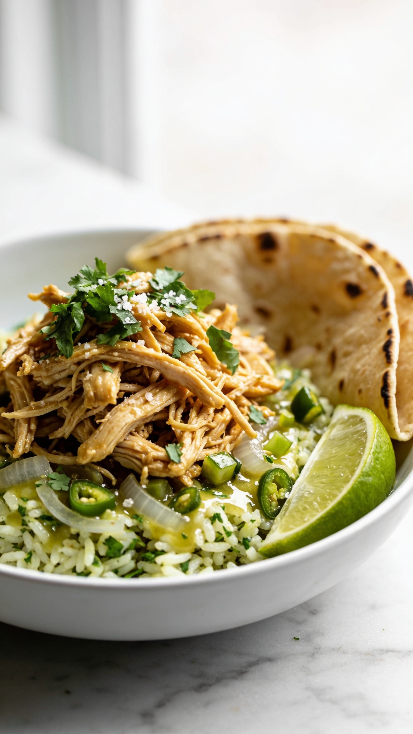 Ultra-close hero shot of High Protein Crockpot Cilantro Lime Chicken: juicy shredded chicken breast piled in a shallow white bowl over fluffy cilantro-lime rice, glistening with a light glossy limey jus; vibrant chopped cilantro scattered throughout, thin lime wedges tucked at the rim, and a few translucent sautĂŠed onions mixed in; tiny diced jalapeĂąo and a sprinkle of coarse sea salt for sparkle; soft charred street-style corn tortillas folded at the edge for serving; bright natural window light from the left, shallow depth of field, creamy bokeh; colors popâgolden chicken shreds, fresh green cilantro, vivid lime; clean marble surface, no text; food fills 90â95% of frame, premium food-blog styling.