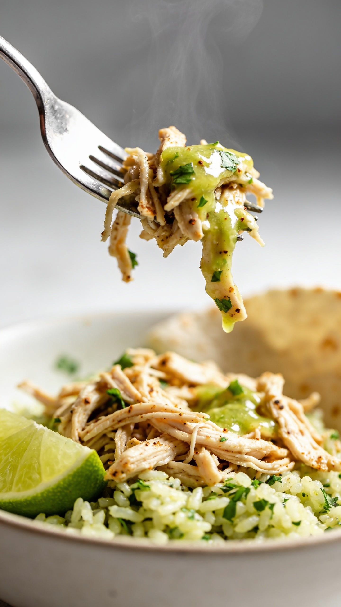 Dynamic action close-up: a polished fork lifting the perfect bite of the same cilantro lime shredded chicken from the bowl, a few strands of juicy chicken draping off the fork with glossy lime-cilantro jus clinging, tiny flecks of cilantro and cumin-chili seasoning visible; a wisp of steam rising; below the fork, the bowl shows the same setupâshredded chicken over cilantro-lime rice, lime wedge, and tortilla edgeâsoftly blurred; bright natural side light, shallow depth of field; movement frozen one second before the bite; food dominates 90â95% of frame, no hands or people.