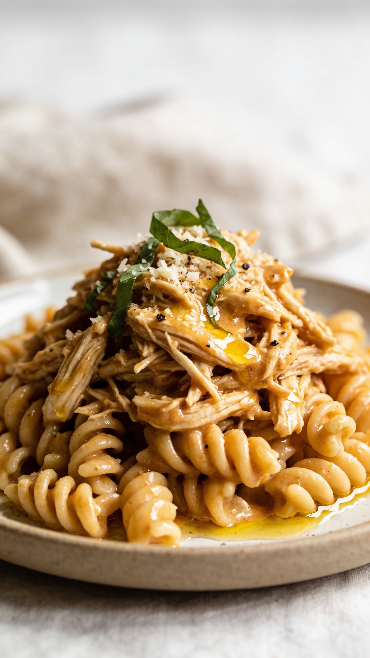 Macro close-up from a low 3/4 angle of the same finished dish: the saucy shredded chicken mounded over rotini with a glossy, creamy sheen, sauce pooling in pasta grooves, edges of chicken fibers glistening; sprinkled Parmesan melting slightly into the warm sauce, basil ribbons and a few cracks of black pepper on top; a drizzle of golden olive oil catching highlights; bright natural light, shallow depth of field focusing on the front mound of chicken and pasta, neutral ceramic plate and soft linen in the background, 90–95% of frame filled with the food, cohesive with the hero and action shots.