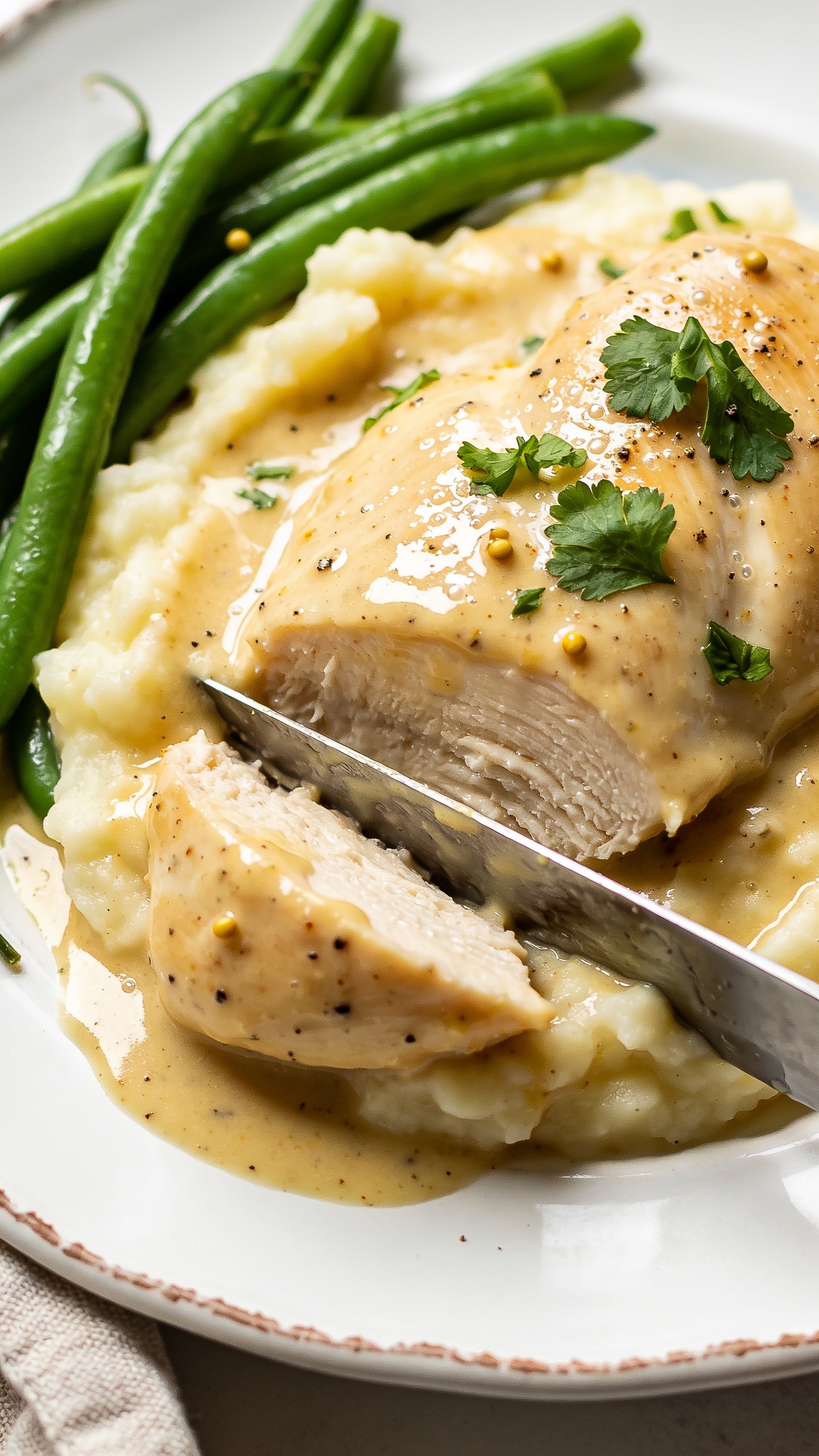 Macro angle from slightly overhead and to the side: the same plated High Protein Crockpot Creamy Dijon Chicken over mashed potatoes, with green beans tucked to the edge. Emphasize juicy textures—knife-cut edges of chicken glistening, creamy Dijon sauce pooled and lightly marbled across the potatoes, micro-beads of moisture catching the light. Fresh parsley and a few mustard seed flecks visible, light pepper dusting. Bright natural light, shallow depth of field isolating the sauced chicken center, subtle linen edge at frame, 90–95% food in frame, cohesive styling with images 1–