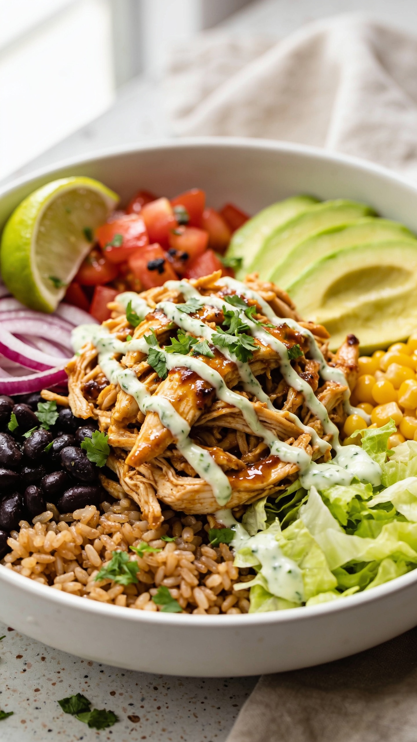 Ultra-close hero shot of a high-protein rotisserie chicken burrito bowl, styled in a matte white shallow bowl, filling 90–95% of the frame. Ingredients arranged in clean, bold layers: cilantro-lime brown rice base, generous mound of shredded rotisserie chicken tossed in glossy chipotle-lime sauce, black beans, charred corn, diced ripe tomatoes, thin-sliced red onion, creamy avocado fan, crunchy romaine, and a drizzle of tangy Greek-yogurt cilantro crema zigzagging over the chicken. Garnish with fresh chopped cilantro and a wedge of lime tucked at the rim. Bright natural window light from the left, soft shadows, shallow depth of field with focus on juicy chicken and crema sheen, background a blurred neutral linen and speckled ceramic surface. Colors vibrant and saturated, textures crisp and glistening, premium food-blog styling.