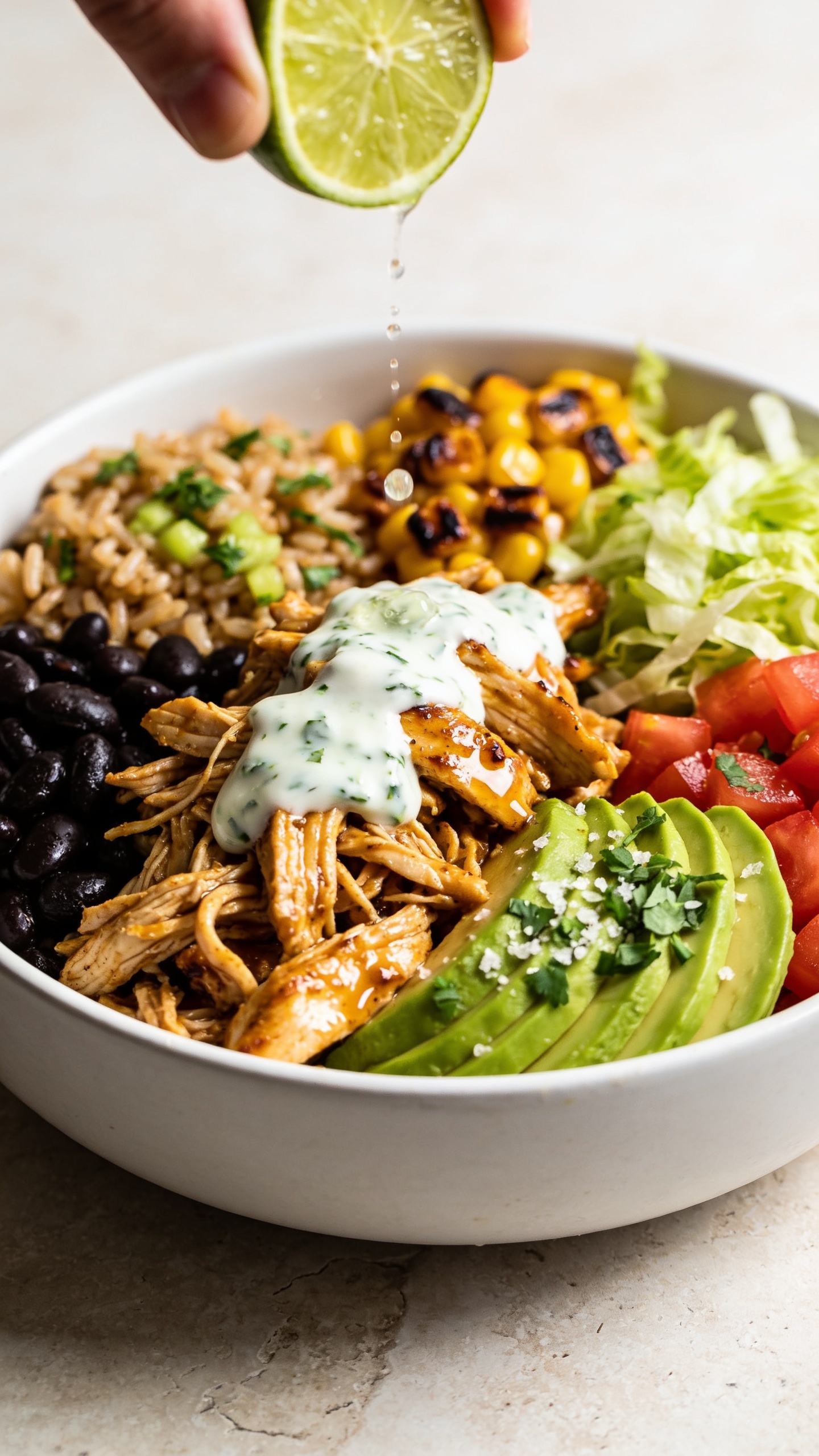 Close-up beauty angle of the same finished burrito bowl from a 45-degree overhead tilt, emphasizing juicy textures: shredded rotisserie chicken glistening under a fresh second drizzle of yogurt-cilantro crema and a light squeeze of lime mid-splash frozen in time, micro-droplets catching the light. Surrounding sections show fluffy cilantro-lime brown rice, glossy black beans, smoky-charred corn kernels, ripe tomato dice, crisp romaine shreds, and velvety avocado fan dusted with flaky salt and chopped cilantro. Bright natural lighting, shallow depth of field focused on the sauced chicken and lime droplets, neutral prop styling (matte white bowl, light stone surface) to keep the food dominant at 90–95% of the frame.