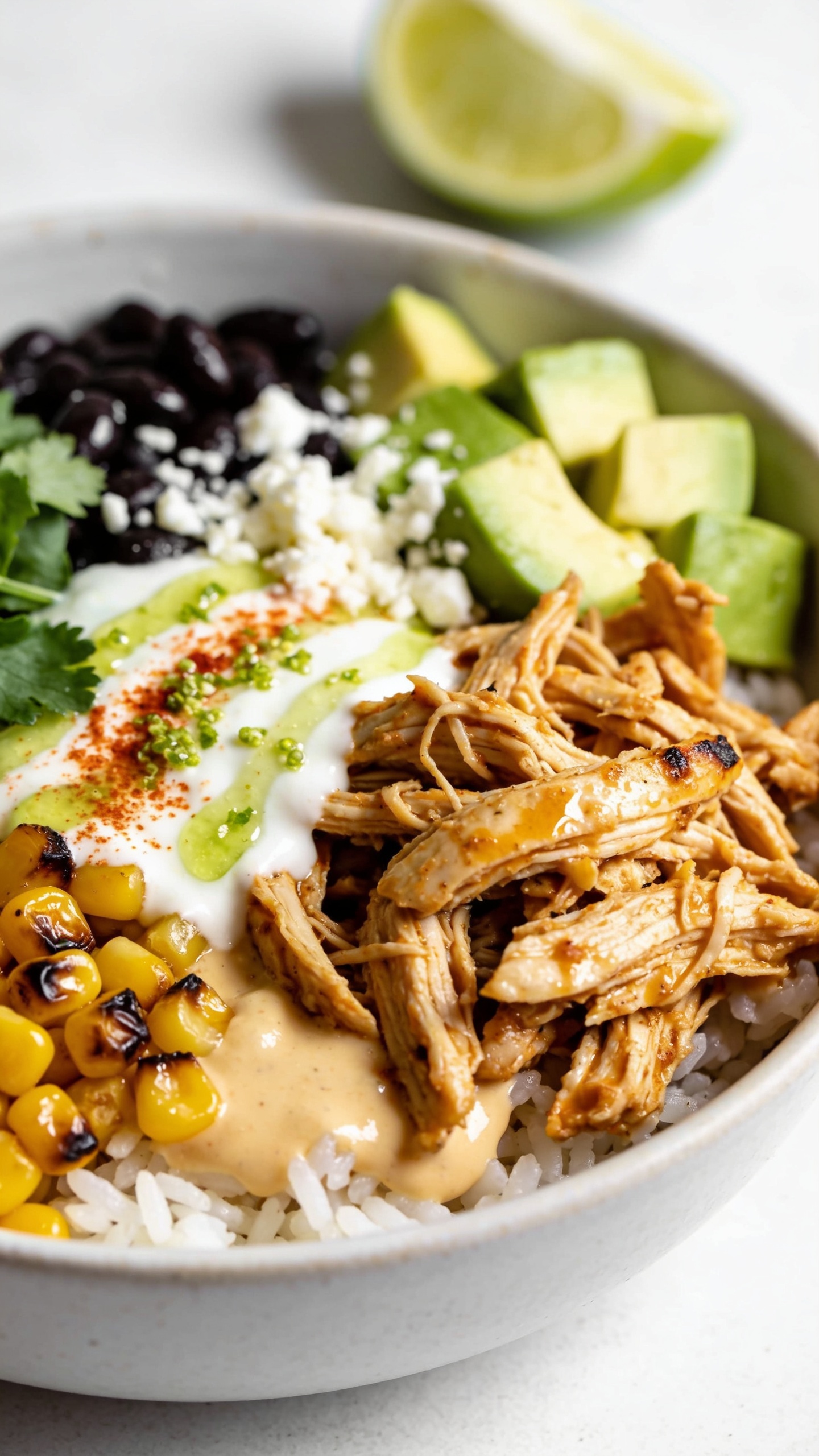 Macro side-angle close-up of the same finished bowl emphasizing textures: focus on the junction where juicy shredded chicken meets charred corn and creamy sauce pooling slightly over fluffy white rice. Visible paprika-speckled yogurt-lime drizzle with micro-beads of lime zest, crumbled cotija, black beans glistening with light, avocado cubes with clean edges, and fresh cilantro leaves. A squeeze-marked lime wedge rests in the background blur. Bright natural daylight, shallow depth of field, crisp highlights on glossy sauce and moist chicken fibers, vibrant colors, premium food-blog styling, no text, no hands, food occupying 90–95% of frame.