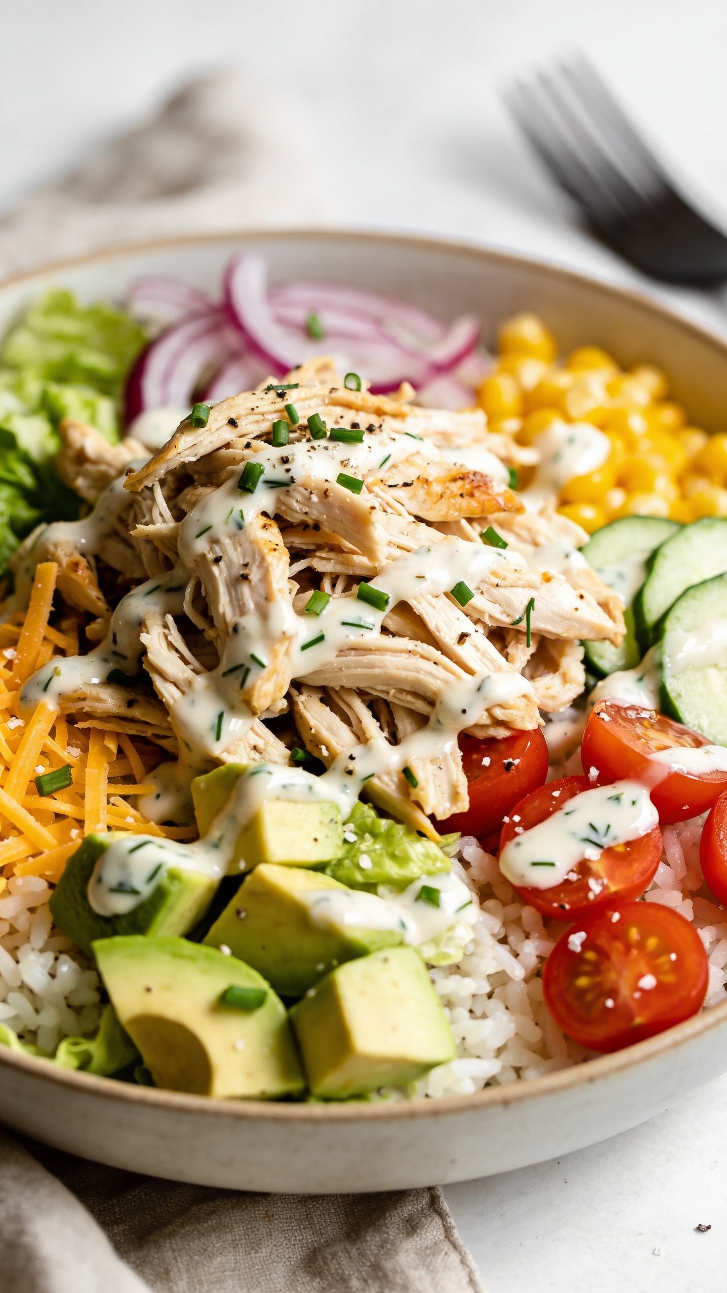 Ultra-close hero shot of a High-Protein Rotisserie Chicken Ranch Bowl: shallow ceramic bowl filled 90–95% of frame with shredded rotisserie chicken piled high over a half-and-half base of fluffy white rice and chopped crisp romaine, drizzled in glossy speckled Greek-yogurt ranch (visible dill and chives), dotted with ripe avocado cubes, cherry tomato halves, thin cucumber slices, shaved red onion, sweet corn kernels, and a sprinkle of shredded sharp cheddar and chopped fresh chives; a light dusting of black pepper and flaky salt; bright natural side lighting, shallow depth of field, soft linen napkin and matte fork blurred in background, colors vibrant and fresh, premium food-blog styling.
