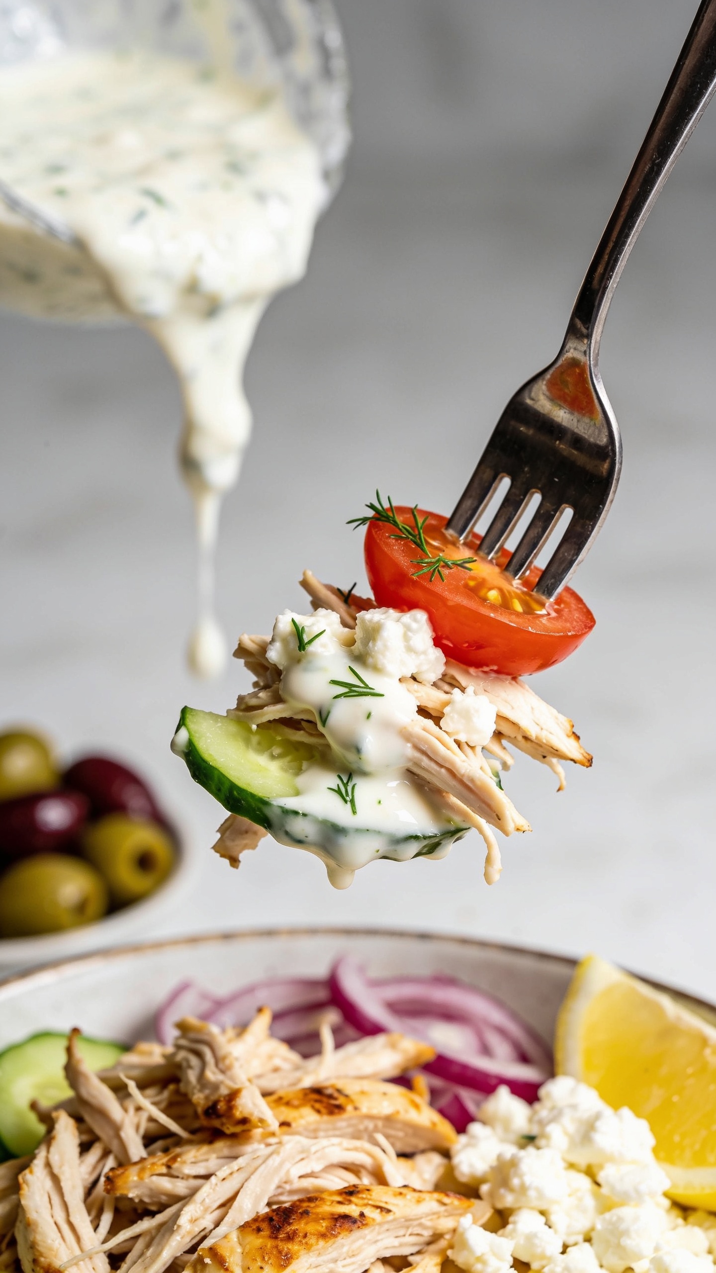 Dynamic action close-up: a polished fork lifting the perfect bite from the same Greek bowl—shredded rotisserie chicken strands topped with glossy tzatziki, a crumble of feta, a sliver of cucumber, and a cherry tomato half speared on the tines, tiny flecks of dill clinging to the sauce. A delicate drizzle of tzatziki is mid-pour in the background, frozen in motion, catching natural light and creating a silky ribbon effect. Background blur reveals hints of olives, red onion, and lemon wedge in the bowl below, all in bright natural lighting. Ultra shallow depth of field, the lifted bite sharp and mouthwatering one second before the bite. Food dominates 90–95% of frame, premium magazine feel, no hands or faces, only the utensil.