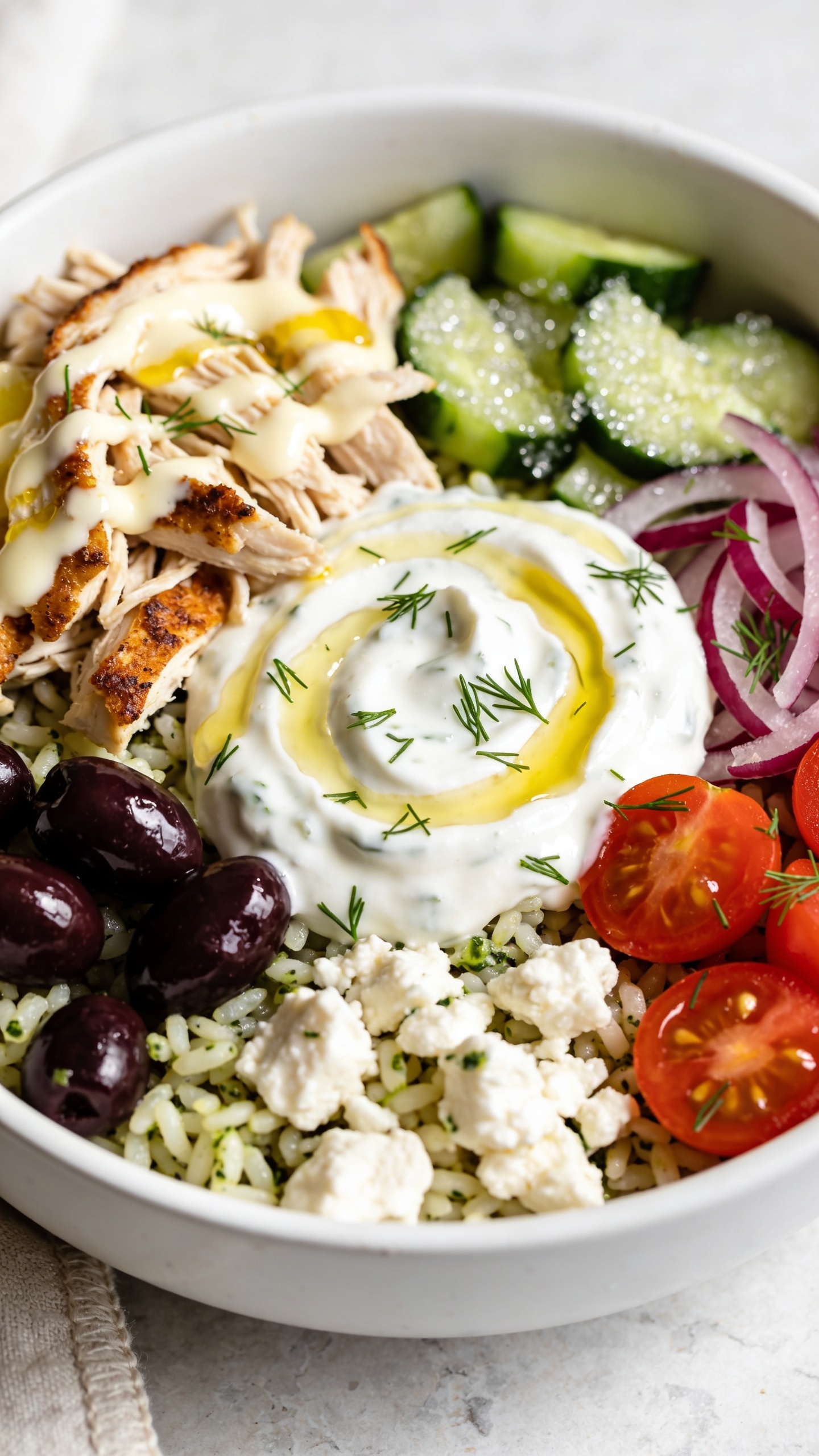 Extreme close-up, angled top-side view of the same finished Greek bowl focusing on juicy shredded chicken nestled over lemon-herb rice with glistening kalamata olives, briny feta crumbles, crisp cucumber, red onion slivers, and cherry tomato halves; a glossy swirl of tzatziki pooled and lightly streaked with extra-virgin olive oil, micro-beads of moisture on the cucumbers, and fresh dill sprinkled like confetti. Bright natural light, shallow depth so textures pop—crispy chicken edges, creamy sauce, grain-by-grain rice definition. White ceramic bowl on a light stone surface with a soft linen edge barely visible. Food fills 90–95% of the frame, cohesive styling with the hero image, no text, no people.