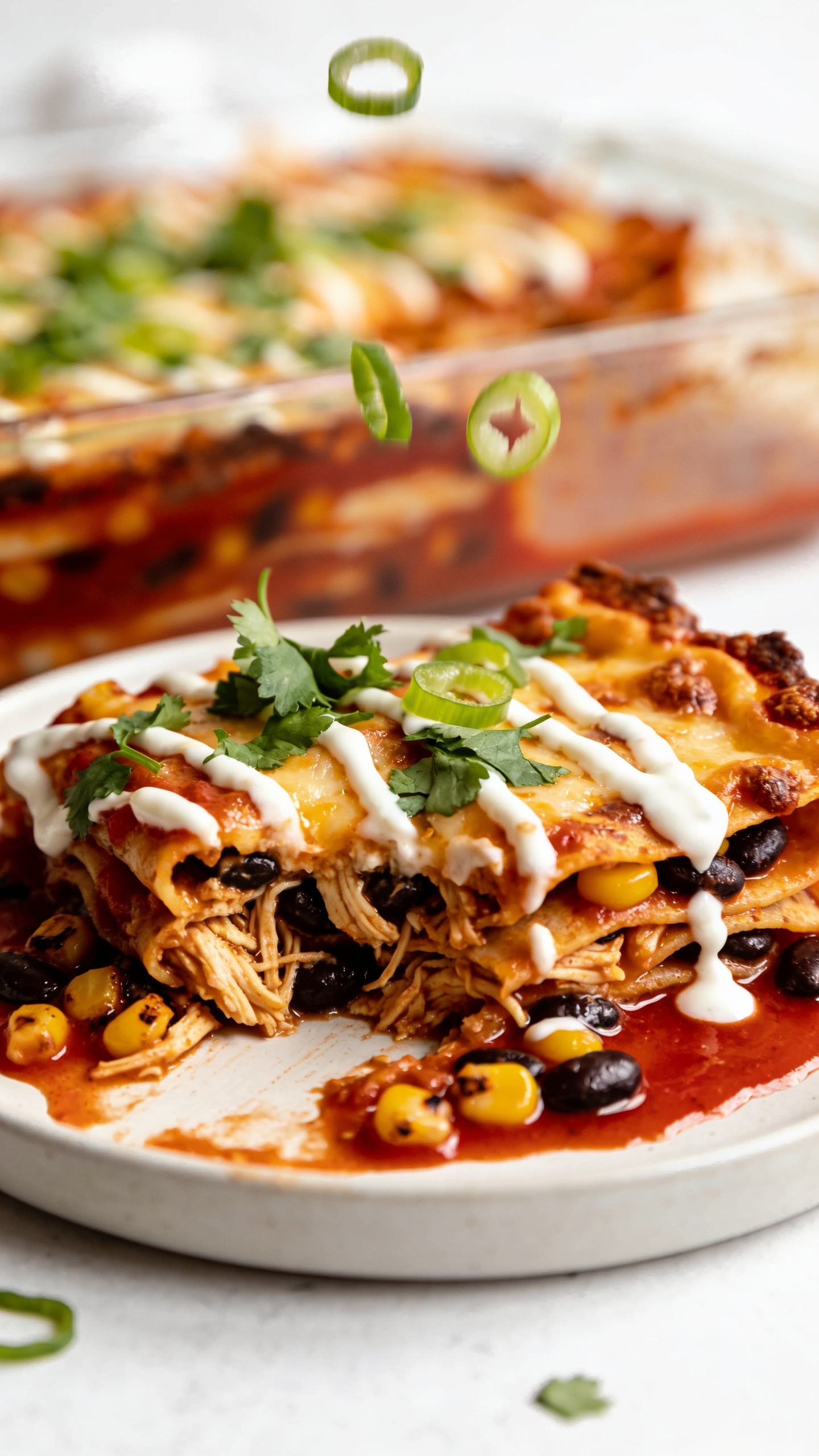 Extreme close-up from a low angle focusing on the just-cut edge of the same casserole slice plated on a matte white plate, showcasing juicy textures: glistening red enchilada sauce pooling slightly at the base, crispy browned cheese edges, tender saucy tortillas, visible chicken fibers, black beans, and charred corn kernels. A delicate drizzle of creamy Greek-yogurt crema zigzags across the top, finishing with a shower of fresh cilantro and a few green onion rings caught mid-fall. Bright natural lighting, shallow depth of field, 90–95% food in frame, cohesive styling matching the baking dish scene.