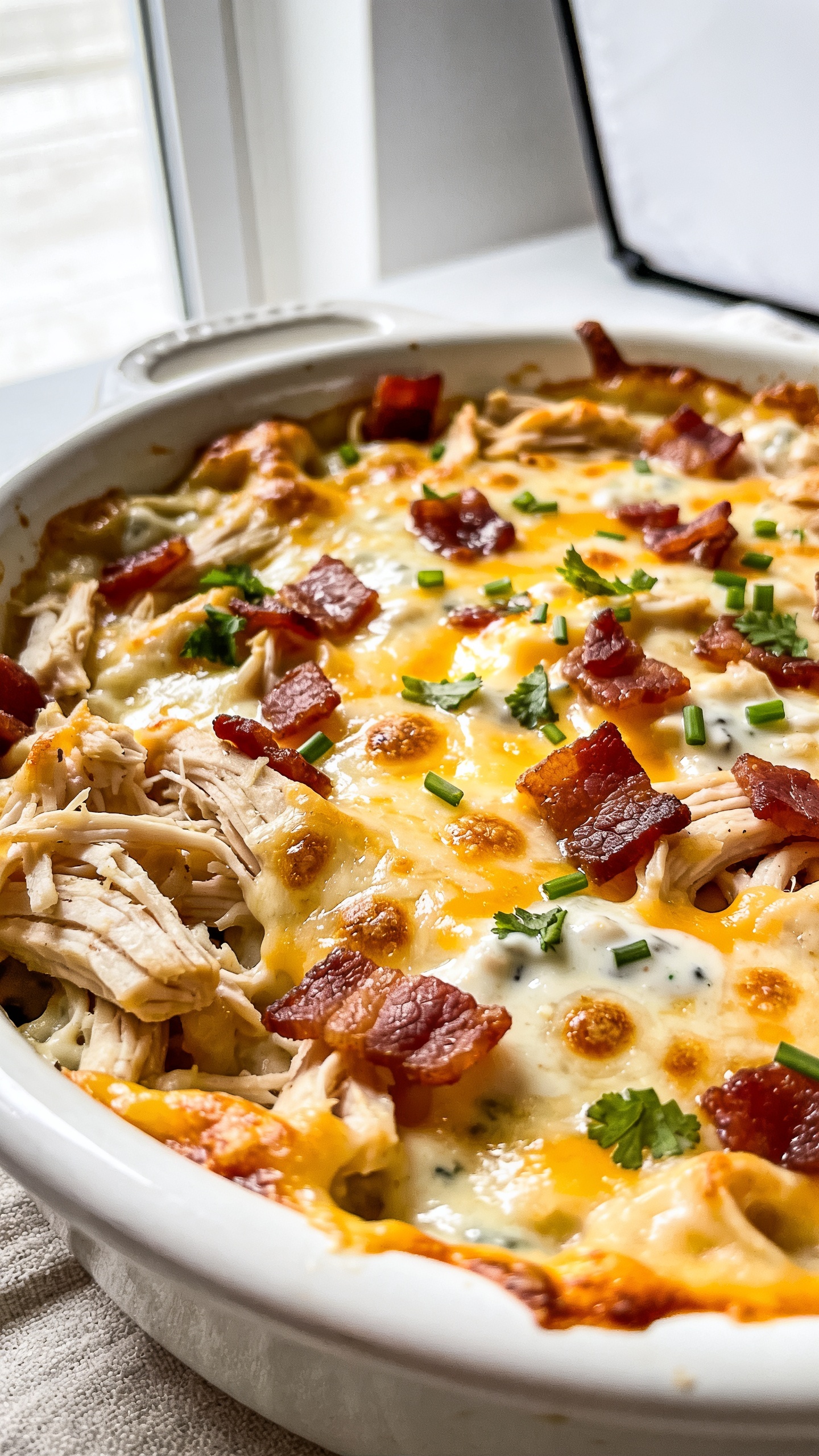 Extreme close-up hero shot of a bubbling, golden-brown High Protein Rotisserie Chicken Bacon Ranch Casserole in a white enamel baking dish, filling 90–95% of the frame. Visible textures: juicy shredded rotisserie chicken, crispy chopped bacon scattered over the top, melted mozzarella/cheddar blend with light brown blistered spots, creamy ranch-infused sauce glossing the surface, and a final sprinkle of fresh chopped chives and parsley. Edges show slightly crispy, caramelized cheese. Bright natural window light from the left, soft reflector on the right, shallow depth of field focusing on the center where cheese is most bubbly. Neutral matte white background and a faint linen hint, premium food-blog styling, no text, no hands.