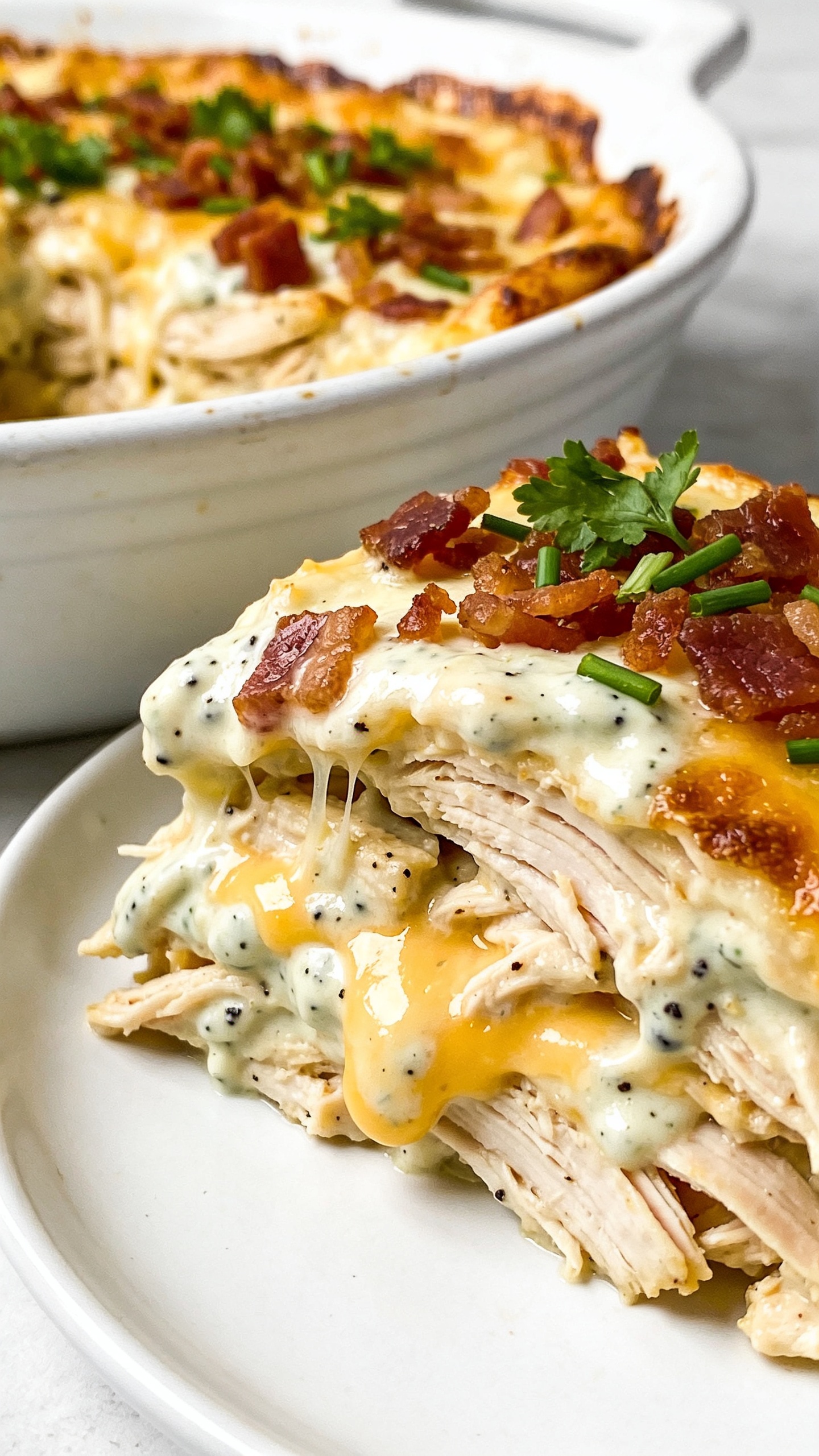 Ultra close-up angled slice view plated from the same casserole on a simple white plate set beside the original white enamel dish in the background, both tightly framed to fill 90–95% of the image. Emphasize the luscious interior: layers of shredded rotisserie chicken coated in creamy ranch sauce, flecks of black pepper, pockets of melted cheese, and crunchy bacon crumbles on top. A light sprinkle of fresh parsley and chives adds vivid green contrast. Slight cheese pull from the plated slice back to the dish, glossy highlights on the sauce, crispy browned edges visible. Bright natural side light, shallow depth of field, premium food-blog look, no text, no hands.