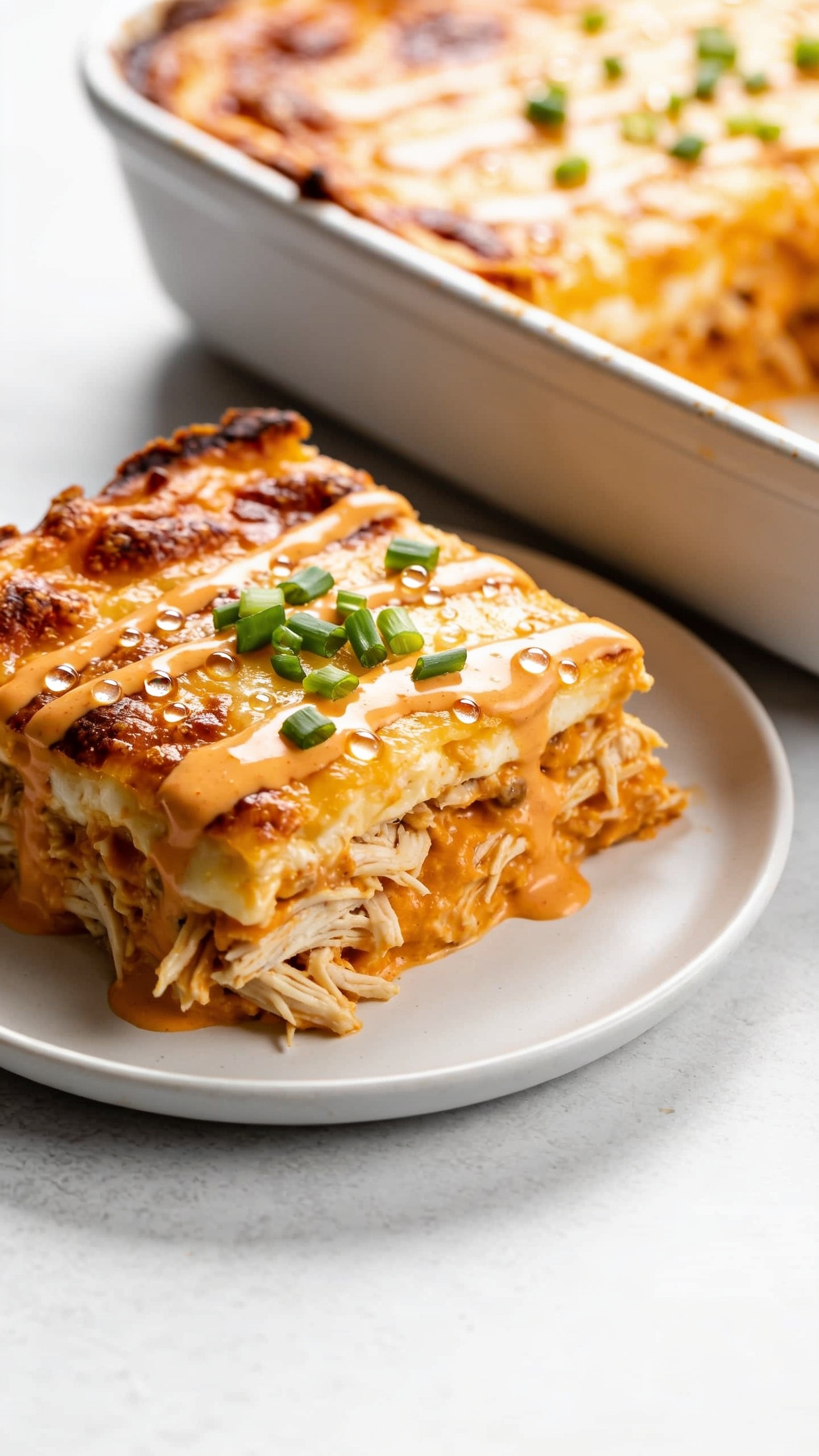 Extreme close-up from a low 3/4 angle of the sliced square of the same casserole plated on a simple white plate placed beside the baking dish, emphasizing layered textures: moist shredded chicken, creamy orange-tinted sauce, and a bronzed, blistered cheesy crust with crisped edges. A light buffalo sauce drizzle glistens on top, micro-beads of oil catching the light, with a sprinkle of finely chopped green onions for fresh pop. Shallow depth of field, bright natural light, minimal neutral props, rich colors and textures filling 90–95% of the frame.