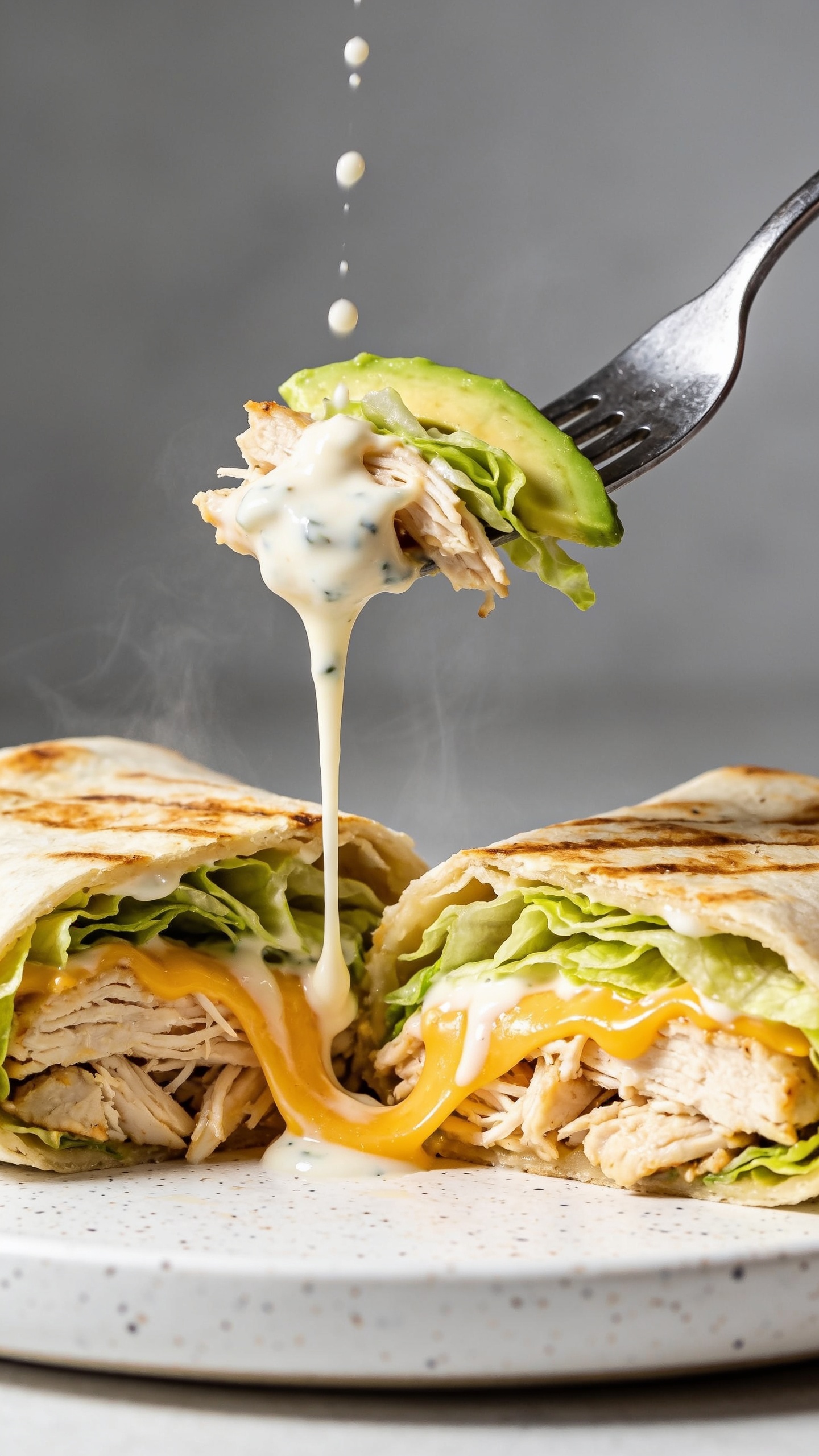 Dynamic action close-up of the same halved rotisserie chicken ranch wrap with a fork lifting the perfect bite from the cut edge—shredded chicken, a ribbon of melted cheddar, a slick of glossy ranch-Greek yogurt sauce stretching slightly, strands of romaine and a sliver of avocado clinging to the fork. A slow drizzle of extra ranch dressing pours in a thin stream onto the exposed filling, captured mid-air as droplets catch the light. No hands visible—only the fork tip and wrap halves on a simple white speckled plate. Bright natural lighting, shallow depth of field, steam faintly visible, food filling 90–95% of the frame, emphasizing juicy textures and creamy gloss.