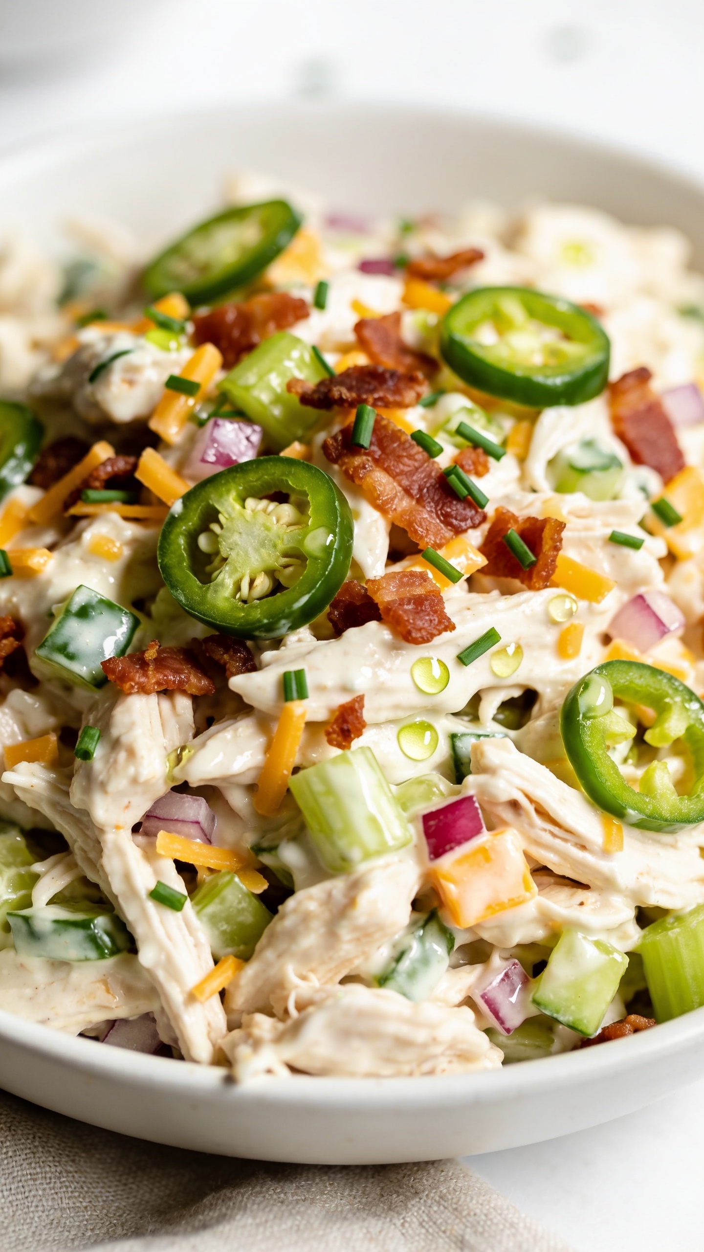 Ultra close-up hero shot of High Protein Jalapeño Popper Chicken Salad in a shallow white bowl, filling 90–95% of the frame. Shredded or chopped roasted chicken bound in a glossy tangy-creamy Greek yogurt dressing, studded with finely chopped fresh jalapeños (visible green flecks and thin rings), crisp celery crescents, tiny red onion dice, and small shards of sharp cheddar melting slightly into the salad. Sprinkled with crumbled crispy turkey bacon and a final scatter of thinly sliced fresh jalapeño and minced chives. A few tiny beads of lime juice glisten on the surface; no visible text. Bright natural side lighting, shallow depth of field, soft neutral linen beneath, clean white background bokeh. Colors vivid: creamy white base, vibrant green jalapeños, golden cheddar, rosy-red onion, and mahogany bacon. Premium food-blog styling, no hands.