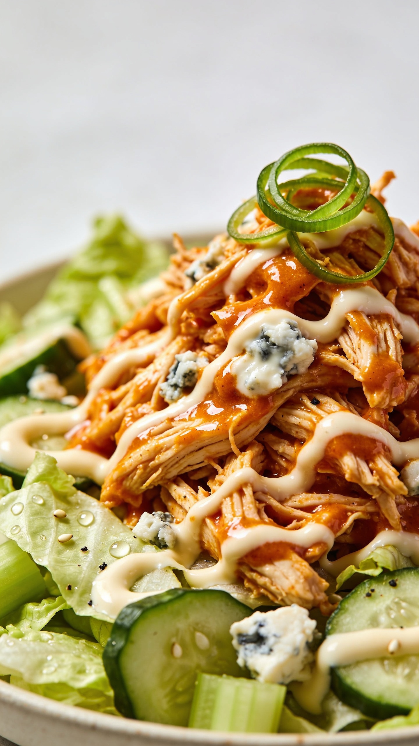 Extreme close-up from a 45-degree angle of the same finished salad just after tossing: shredded rotisserie chicken lacquered in vibrant buffalo sauce nestled into chopped romaine, cucumber, and celery. Visible creamy high-protein dressing marbled through the chicken with glossy streaks, tiny blue cheese crumbles melting slightly into the warm sauce, and a sprinkle of thin green onion rings on top. Emphasize juicy textures: glistening sauce on chicken fibers, crisp edges on cucumbers, beaded moisture on lettuce, and a few sesame-like specks of black pepper. Bright natural side light, shallow depth of field with focus on the sauciest chicken mound and creamy swirls, neutral minimal background to keep the salad dominant in 90–95% of the frame. No text, no hands, no people.