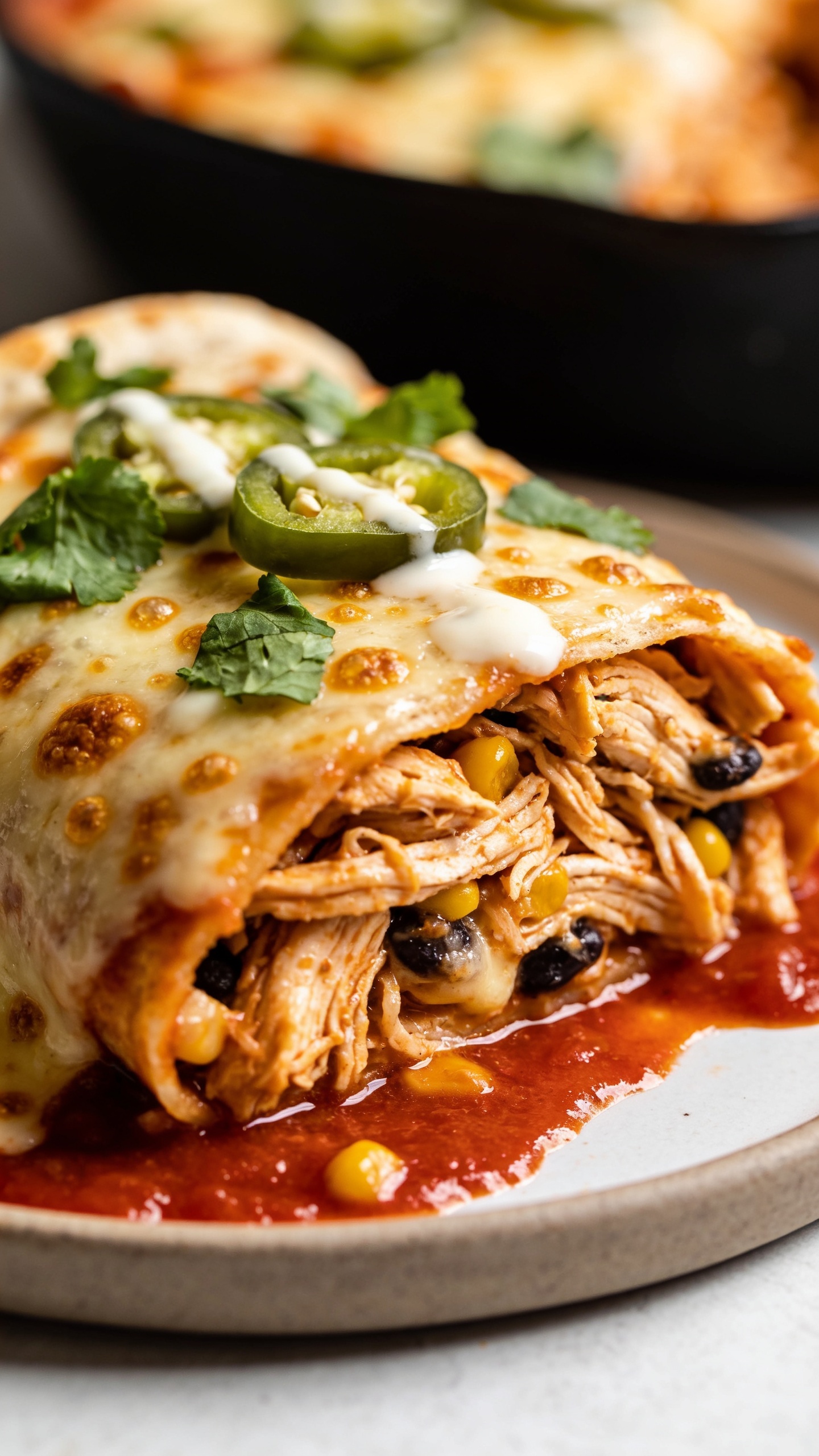 Macro angled close-up of a single sliced enchilada on a small plate pulled from the same pan, revealing the juicy shredded rotisserie chicken filling, specks of black beans or corn tucked among the chicken, edges of tortilla slightly crisp; a rich lacquer of red sauce pooling at the base, cheese molten and lightly browned with bubbly pockets; fresh cilantro scattered on top, a few jalapeño slices and a delicate crema drizzle; vibrant colors, glossy textures, shallow depth of field, bright natural lighting, food filling 90–95% of frame, matching styling to the hero image.