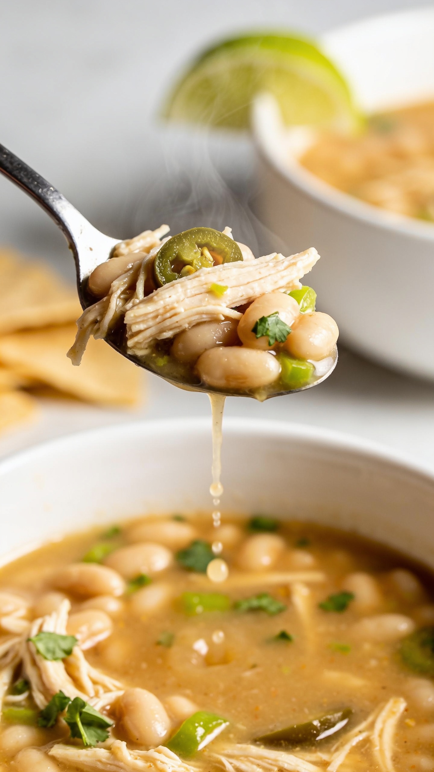 Dynamic action close-up capturing a spoon lifting the perfect, steaming bite of the same white chicken chili one inch above the bowl—spoon brimming with shredded chicken, creamy white beans, flecks of green chile, and a glossy broth thread trailing back into the bowl; micro-beads of broth clinging to the spoon; a few cilantro bits and a tiny jalapeño slice nestled in the scoop; background softly blurred but consistent with the hero setup (white bowl, lime wedge on rim, tortilla strips on the side); bright natural light, shallow depth of field, movement frozen at the most irresistible moment, food filling 90–95% of the frame, no hands visible.