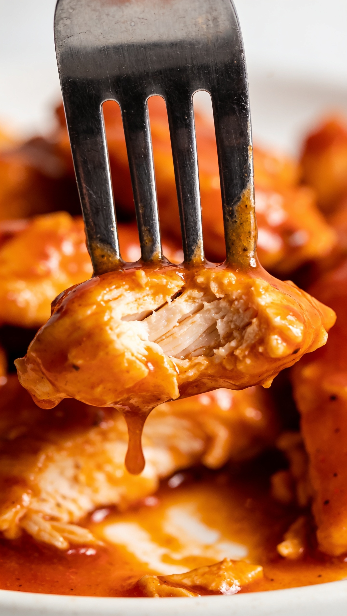 A dynamic close-up action moment: a fork lifting the perfect bite of buffalo chicken from the plate, with the glossy buffalo sauce clinging to the fork tines and a visible juicy interior. The shot is ultra-close, 90–95% food filling the frame, with dramatic shallow depth of field, vibrant colors, and natural lighting; no hands or faces visible, only the bite and sauce captured at the moment of lift.