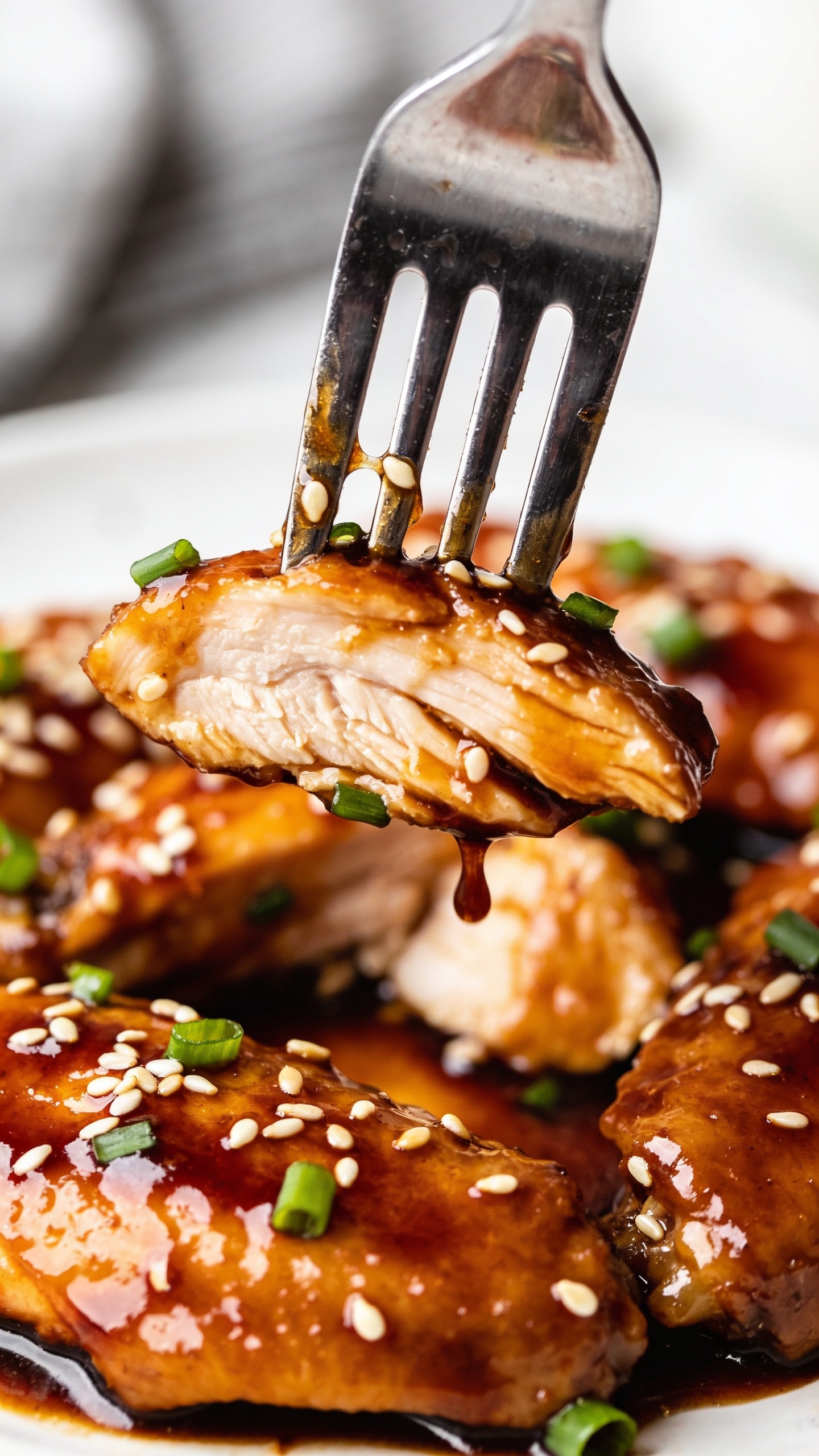 A dynamic, mouthwatering action shot: a fork lifts a perfect bite of the finished chicken from the plate, glaze clinging to the tines and satin gloss visible as it stretches slightly, sesame seeds and green onion flakes catching the light. The image is ultra-close, 90–95% food, bright natural lighting, shallow depth of field, focus on the glossy sauce and juicy interior, hands/arms not visible.