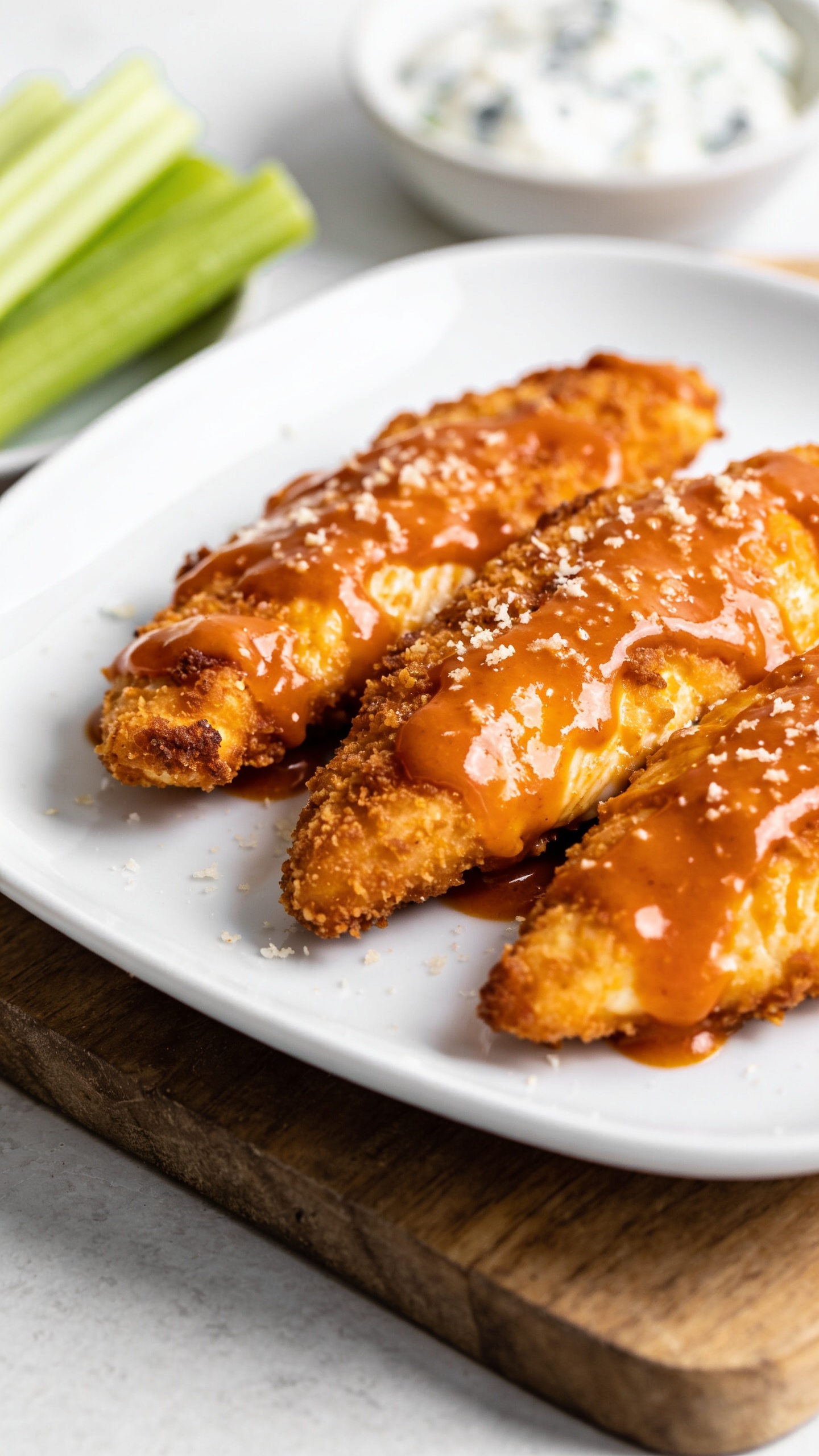 A stunning hero image of the finished High Protein Air Fryer Buffalo Chicken Tenders plated on a sleek white plate with a glossy buffalo glaze, crispy golden-brown crust, and a light dusting of grated Parmesan; the plate sits on a wooden board with a subtle blur of fresh celery sticks and blue cheese dip in the background, natural bright lighting, ultra-close-up, no text, 90–95% food fill.