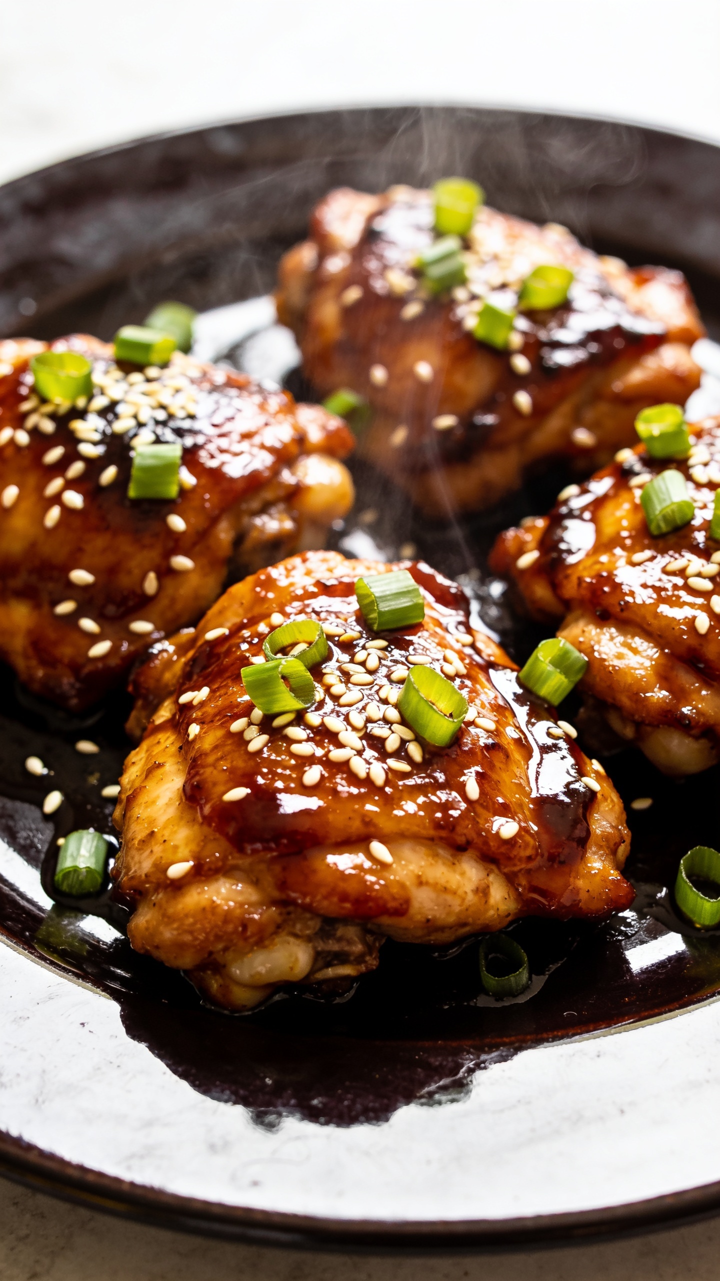 A stunning hero shot of the finished High Protein Sticky Asian Chicken Thighs plated on a glossy, dark plate. The chicken thighs gleam with a thick, caramelized glaze, sesame seeds and sliced green onions sprinkled on top, steam rising subtly. No hands or utensils in frame. Bright natural light, ultra-close-up, shallow depth of field, rich textures and vibrant colors, food filling 90–95% of frame.