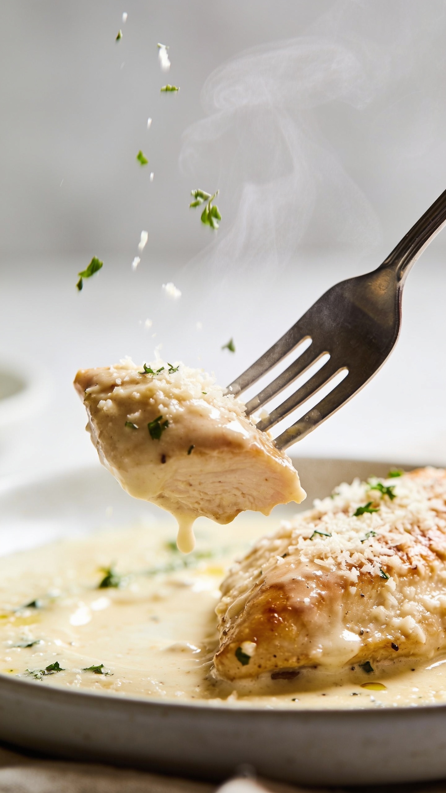 A dynamic close-up action moment: a fork lifts the perfect bite from the plate, the creamy parmesan sauce glistening on the fork and chicken thigh, steam just rising, herbs lightly falling onto the dish, ultra-close with a shallow depth of field, natural bright lighting, no hands visible, 1:1 crop.