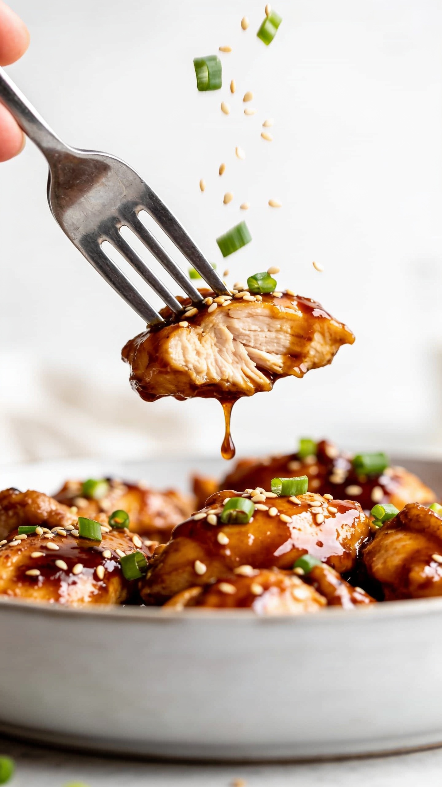 A dynamic close-up action scene of a fork lifting a perfect bite from the finished dish; the fork is hovering just above the plate with glossy bang bang sauce dripping slowly back, the juicy chicken thigh meat visible, sesame seeds and chopped green onions in mid-air as if about to fall, bright natural lighting, ultra shallow depth of field, hands and fingers not visible, the bite filling the foreground.